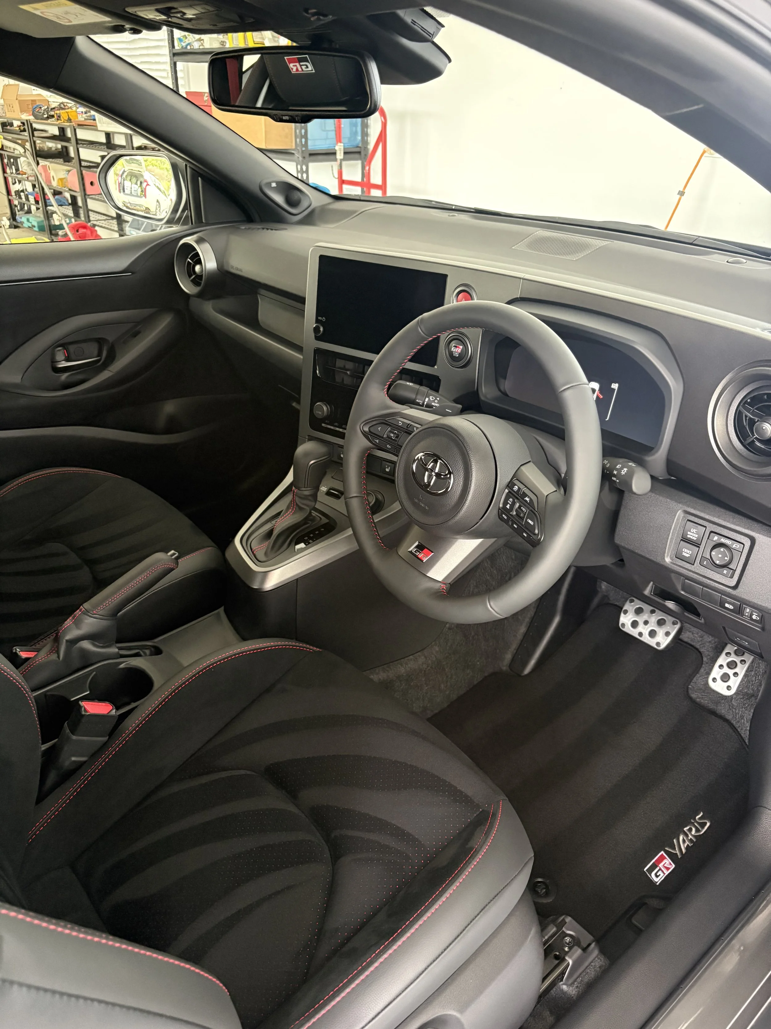 Interior of a Toyota GR Yaris sports car, showing the steering wheel, dashboard, center console, and black seat with red stitching.