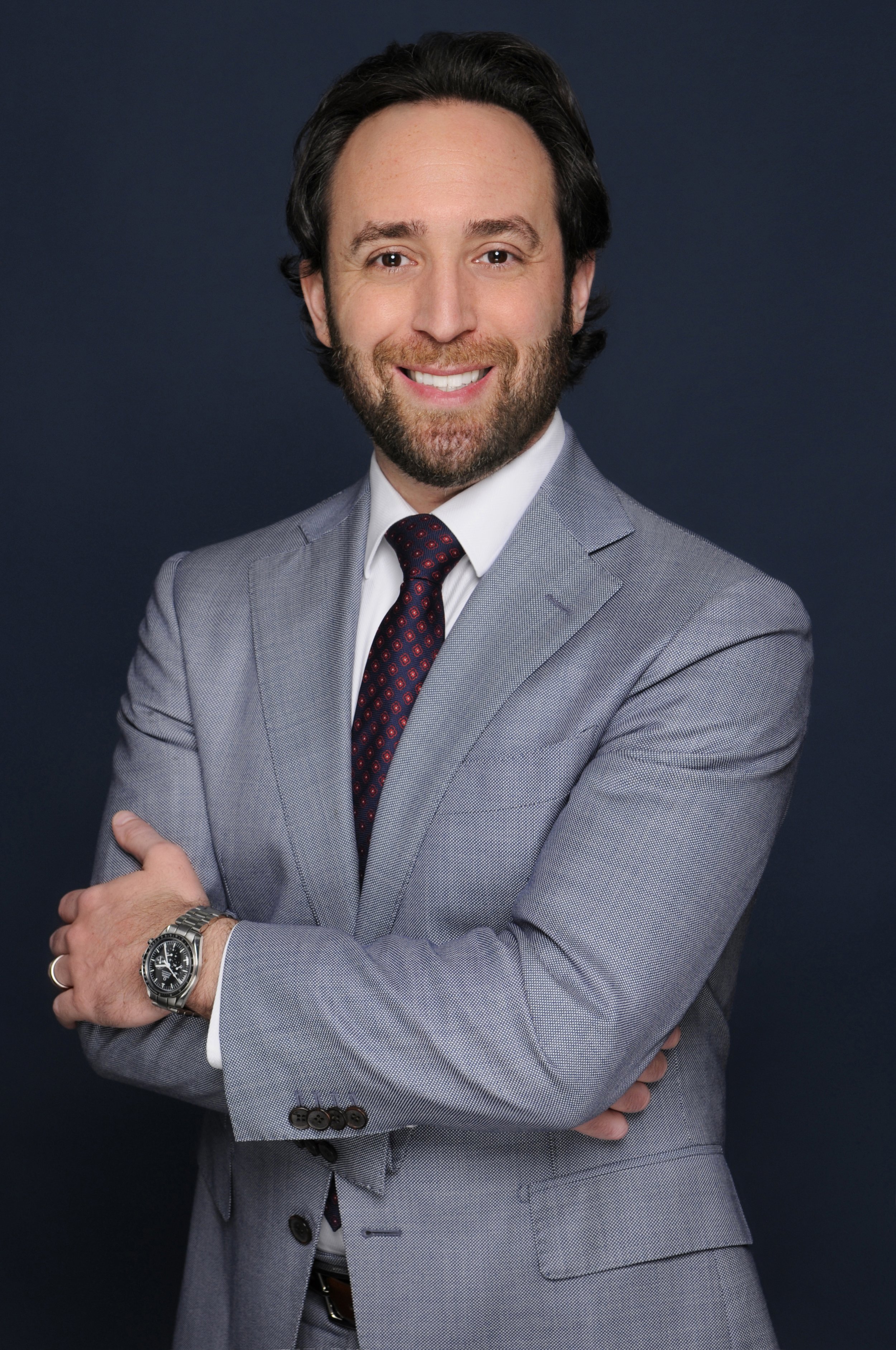 A professional man with dark hair and a beard wearing a gray suit, white shirt, and a red and blue patterned tie, smiling at the camera, with his arms crossed and a watch on his left wrist.