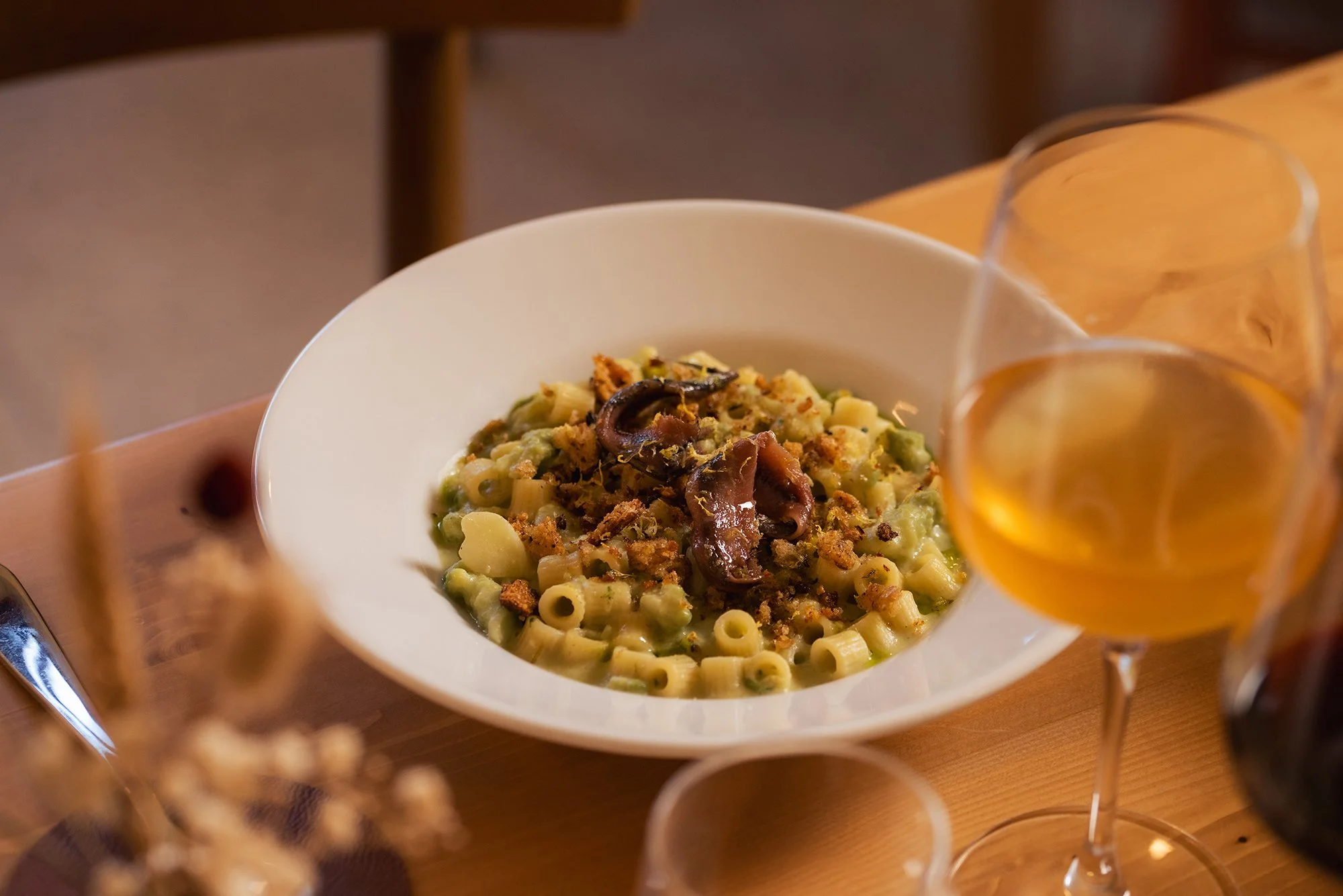 Piatto di pasta con sugo di carne e verdure, accompagnato da un bicchiere di vino bianco, su una tavola di legno.