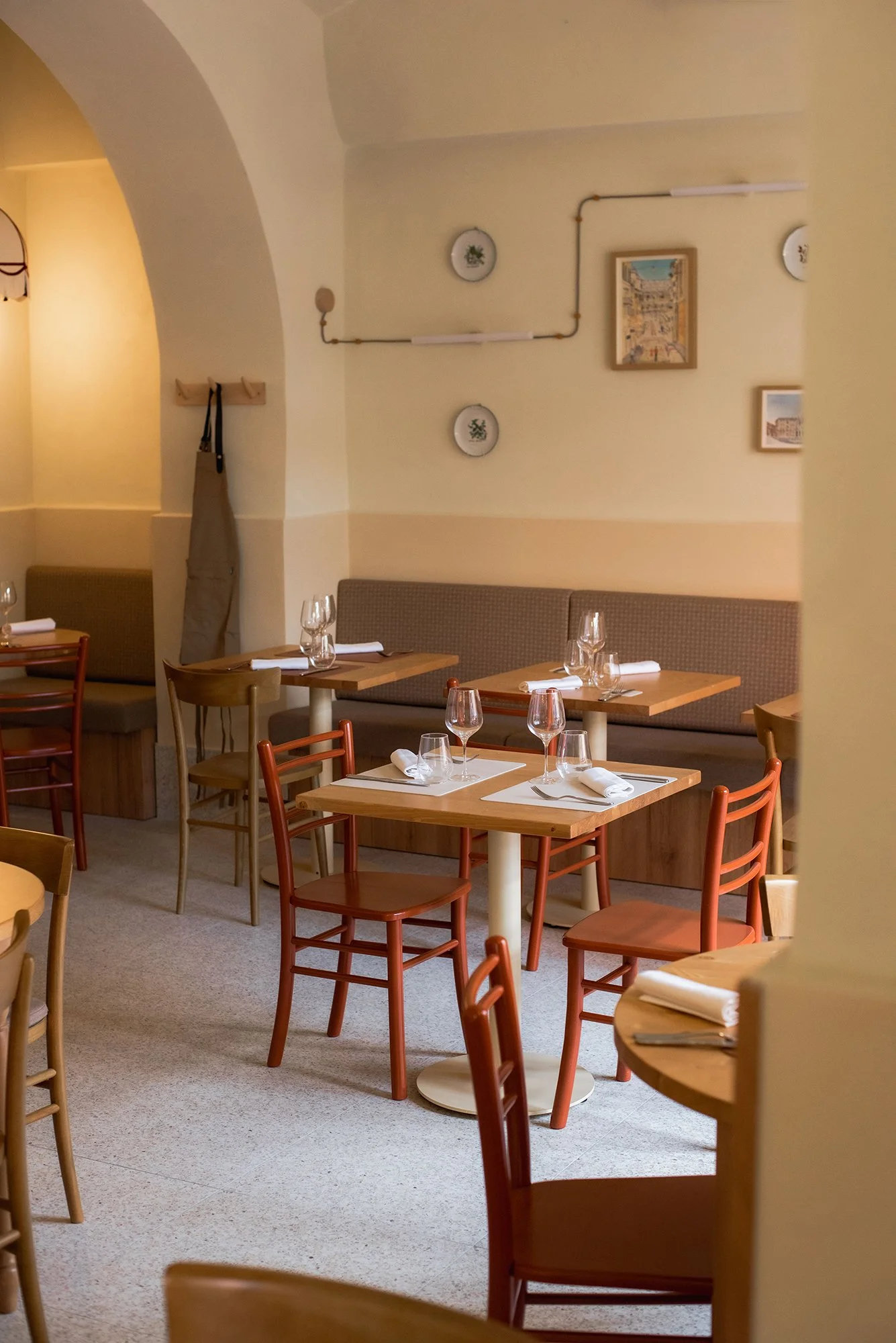 Interno di un ristorante con tavoli in legno, sedie di legno con schienale rosso, apparecchiati con tovagliette bianche, forchette, coltelli, bicchieri da vino e menù. Pareti beige con decorazioni e quadri, e un'area con divano beige.