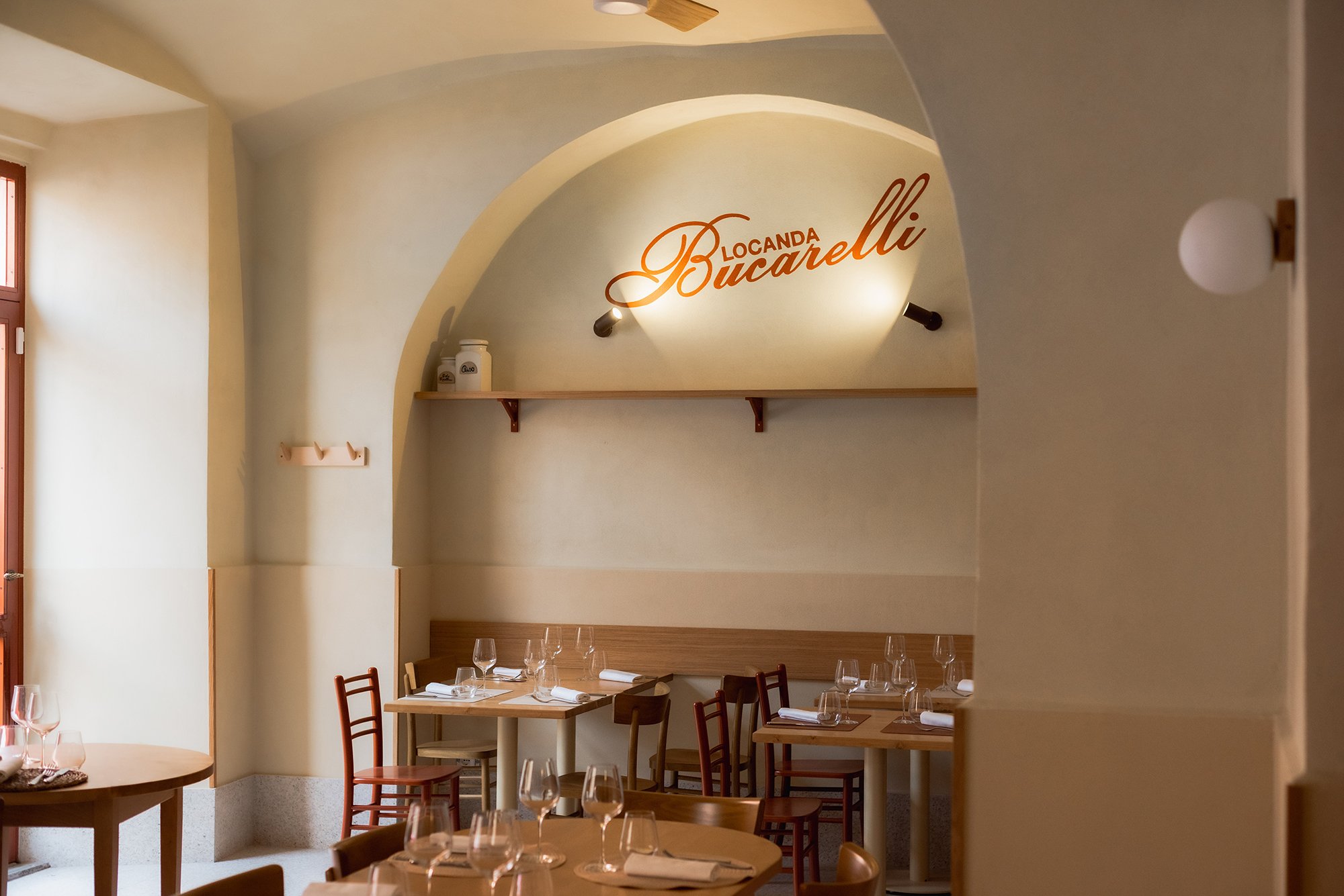Interno di un ristorante con pareti chiare, archetto decorativo con il nome 'Locanda Bucarelli' illuminato, tavoli apparecchiati con calici di vino e posate.