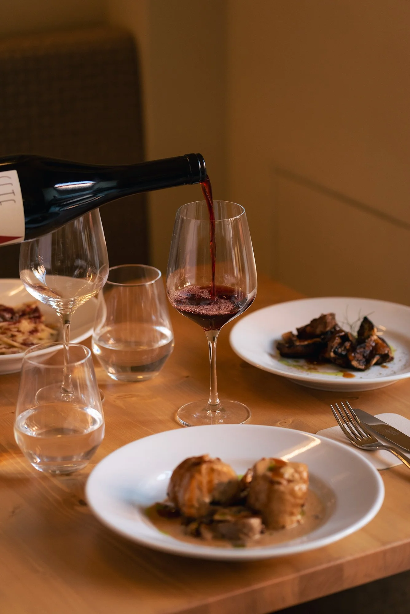 Bottiglia di vino rosso versa in un calice, con altri bicchieri e piatti di cibo sul tavolo.