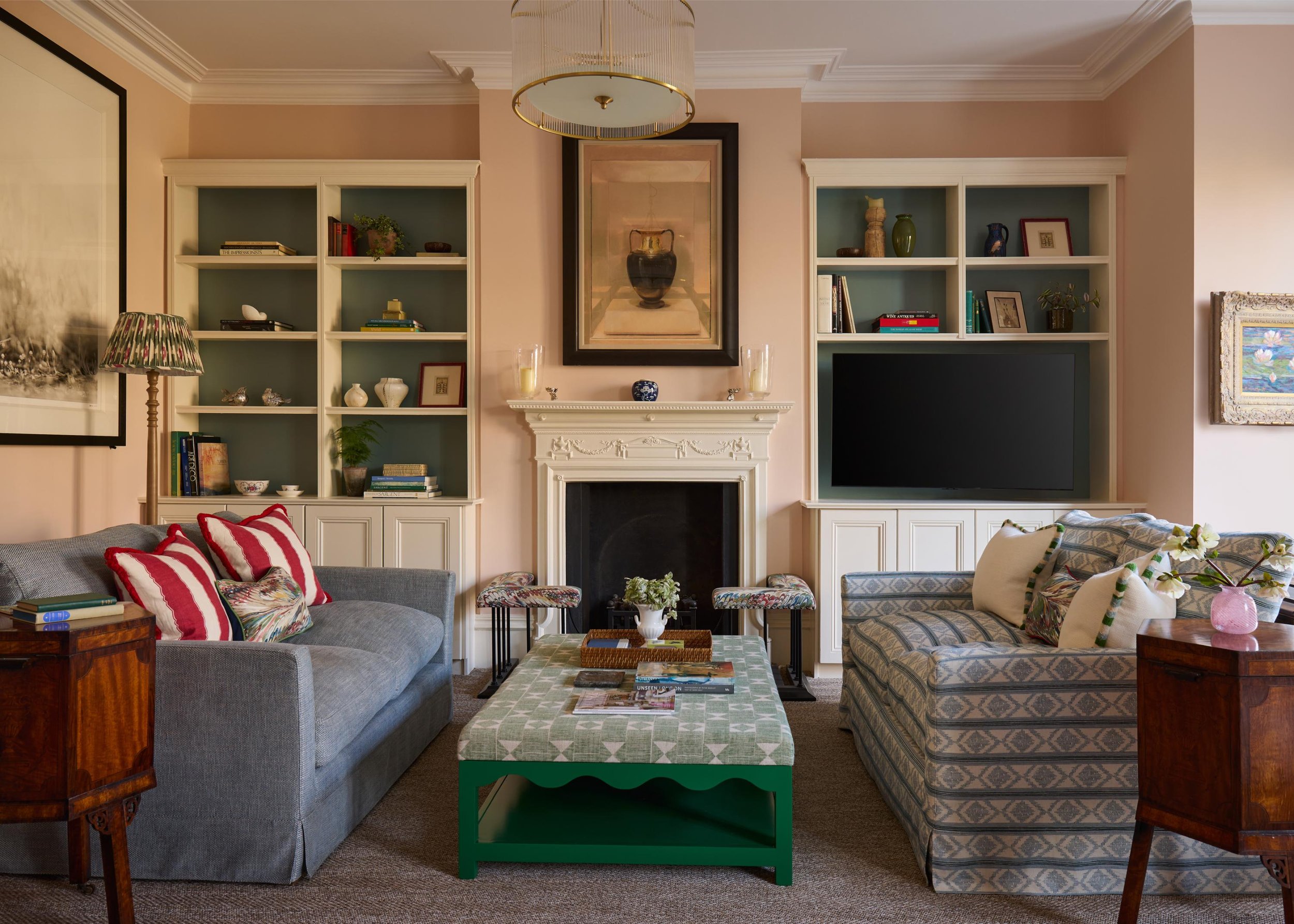 Living room with beige walls, white crown molding, and a fireplace with decorative molding. Two white bookshelves flank the fireplace, filled with books and decorative items. A large framed mirror hangs above the fireplace. There is a gray sofa with 