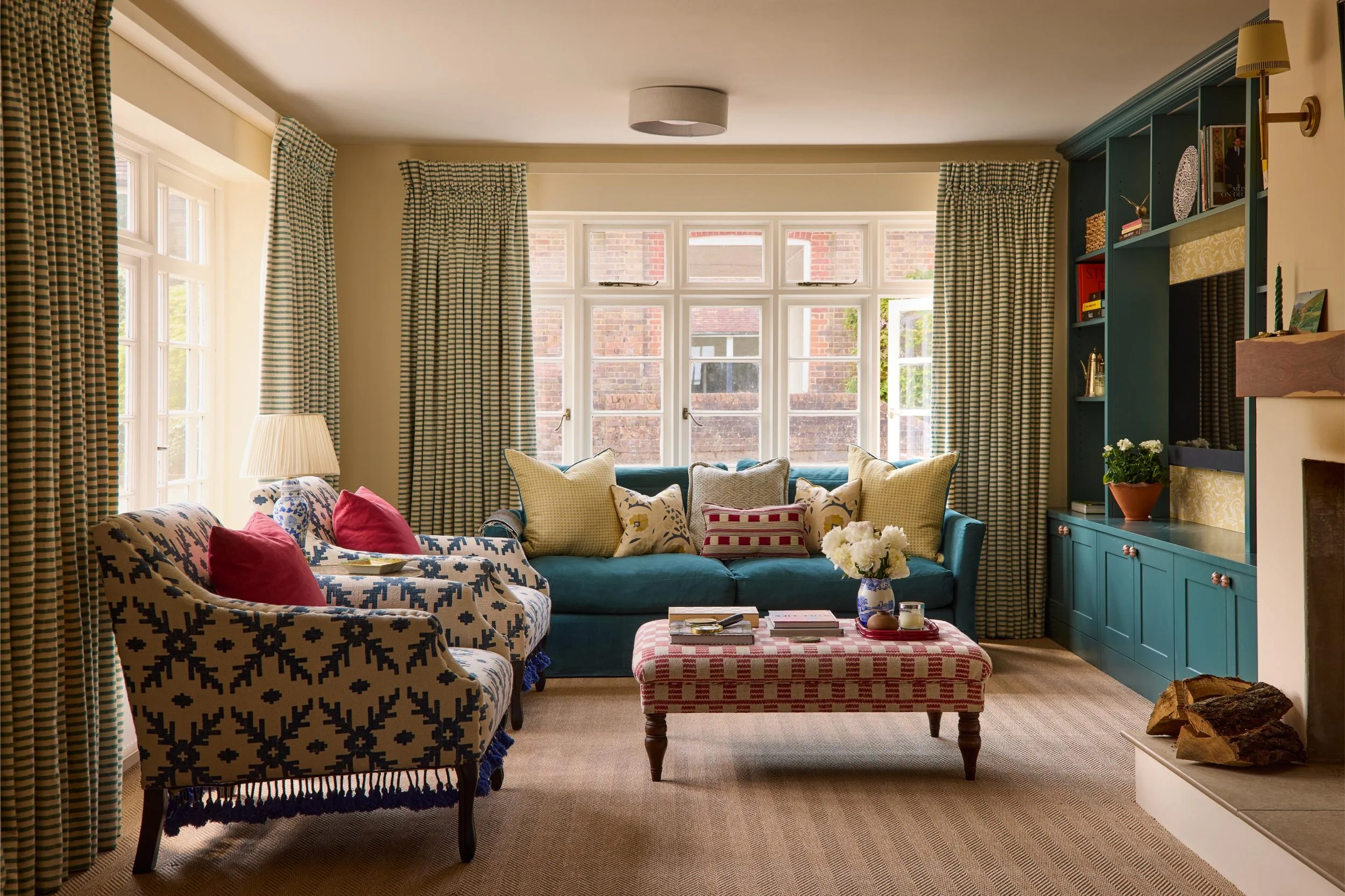 Living room with large windows, cream walls, patterned and solid color sofas with multiple throw pillows, a checkered ottoman, curtains with a blue and white checkered pattern, a blue cabinet with decor and books, a lampshade, a fireplace with wood l