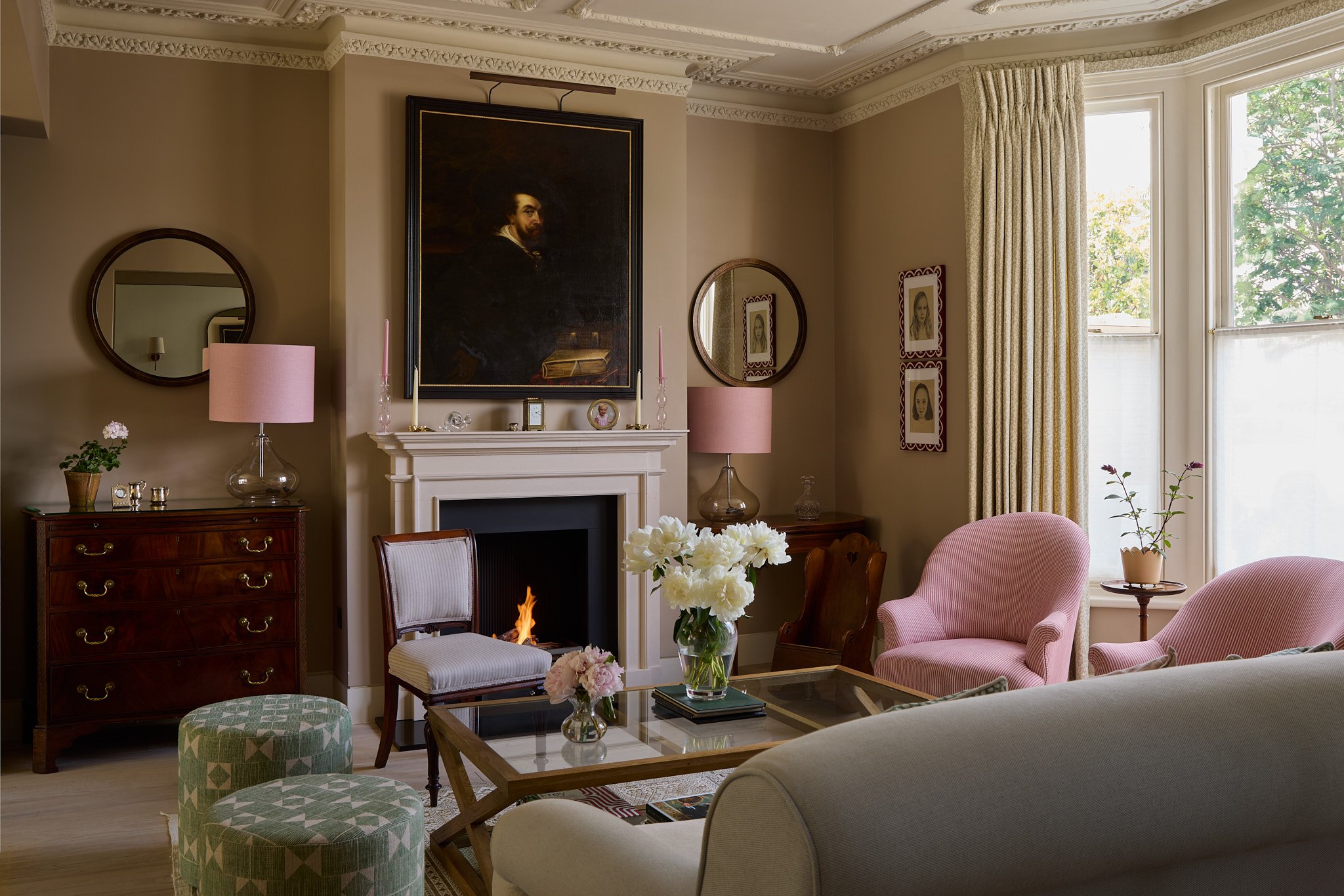 Living room with a fireplace, pink armchairs, and a coffee table with flowers. Large bay window with curtains, framed art, and mirrors on the walls.