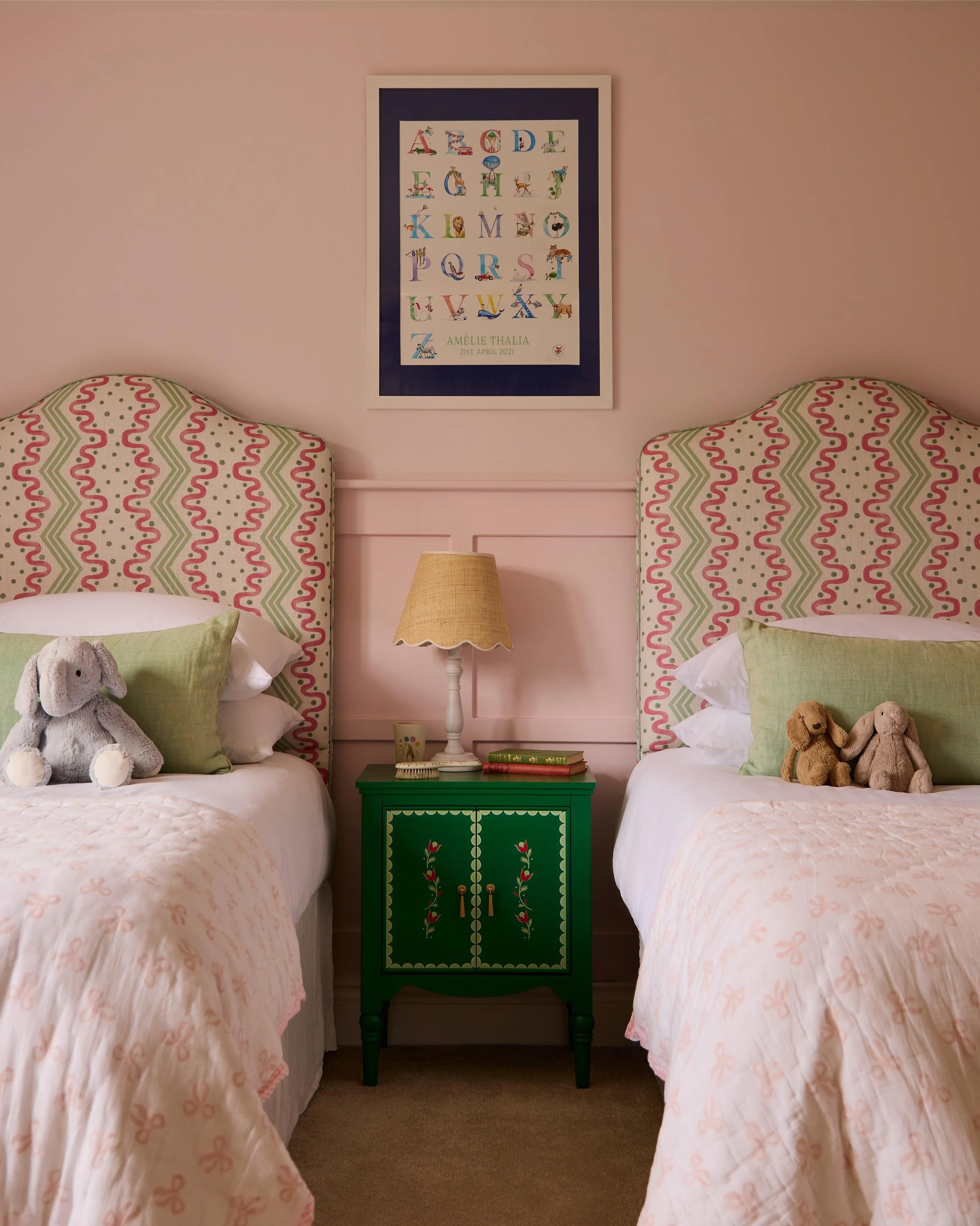 Two beds with pink and white bedding, green ribbon and bow pattern, and plush toys, separated by a green nightstand with decorative floral designs and a wooden lamp. The bedroom has light pink walls, and a framed alphabet print with animal illustrations is hanging above the beds.