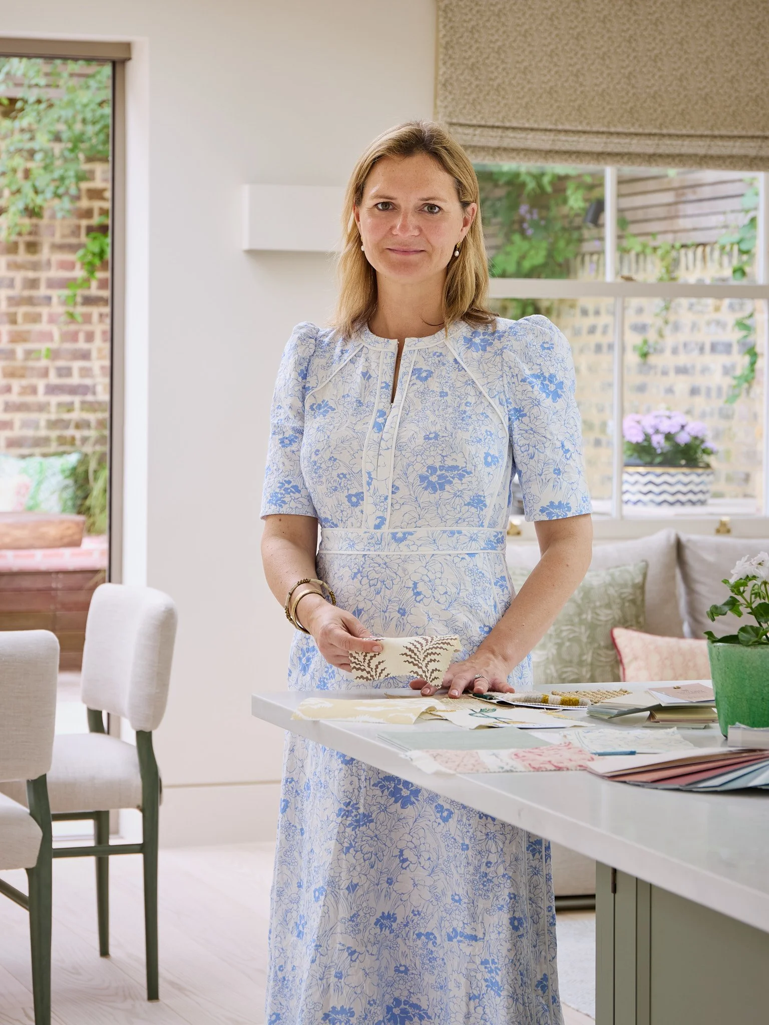 Woman in a light blue floral dress standing at a table with fabric samples, in a bright room with large windows and outdoor garden views.