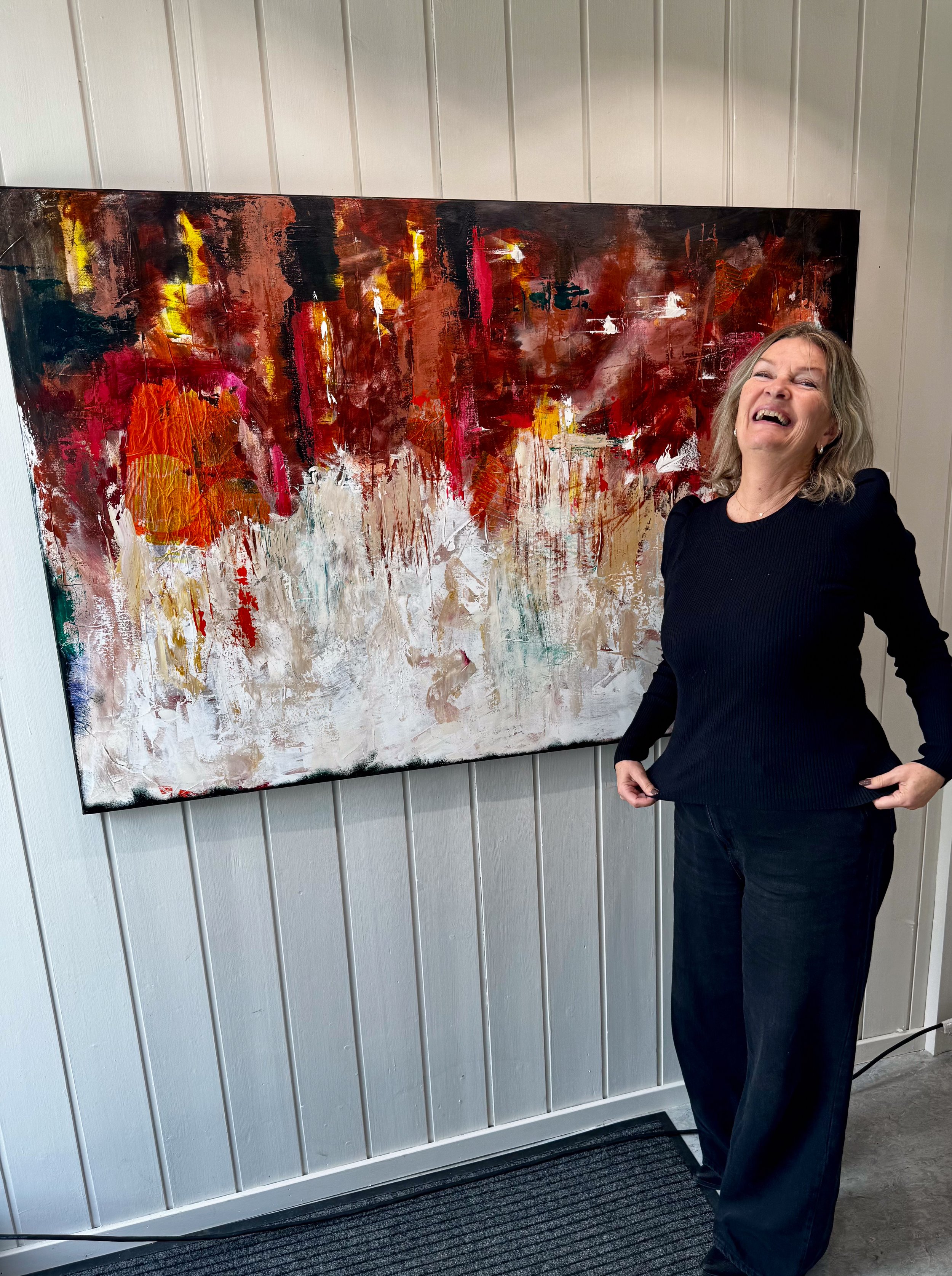 Kunstner Pia C. Bye smiler ved siden av et stort abstrakt maleri i rødt, oransje og hvitt i atelieret