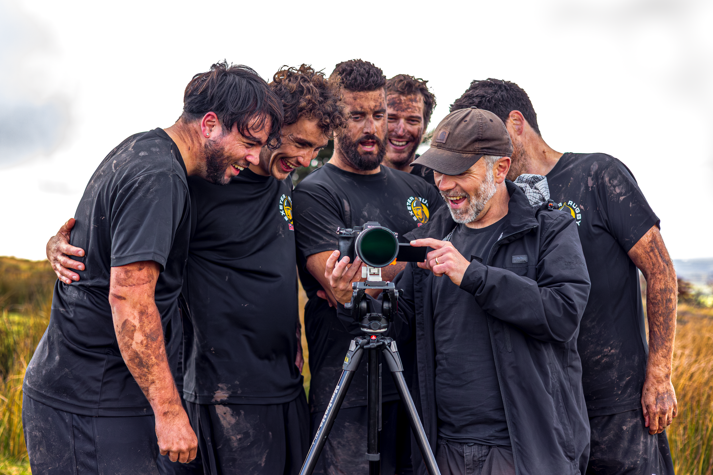 A group of six men, some muddy, are gathered around a camera outdoors, smiling and looking at the camera or screen, likely during a sports event or photo shoot.