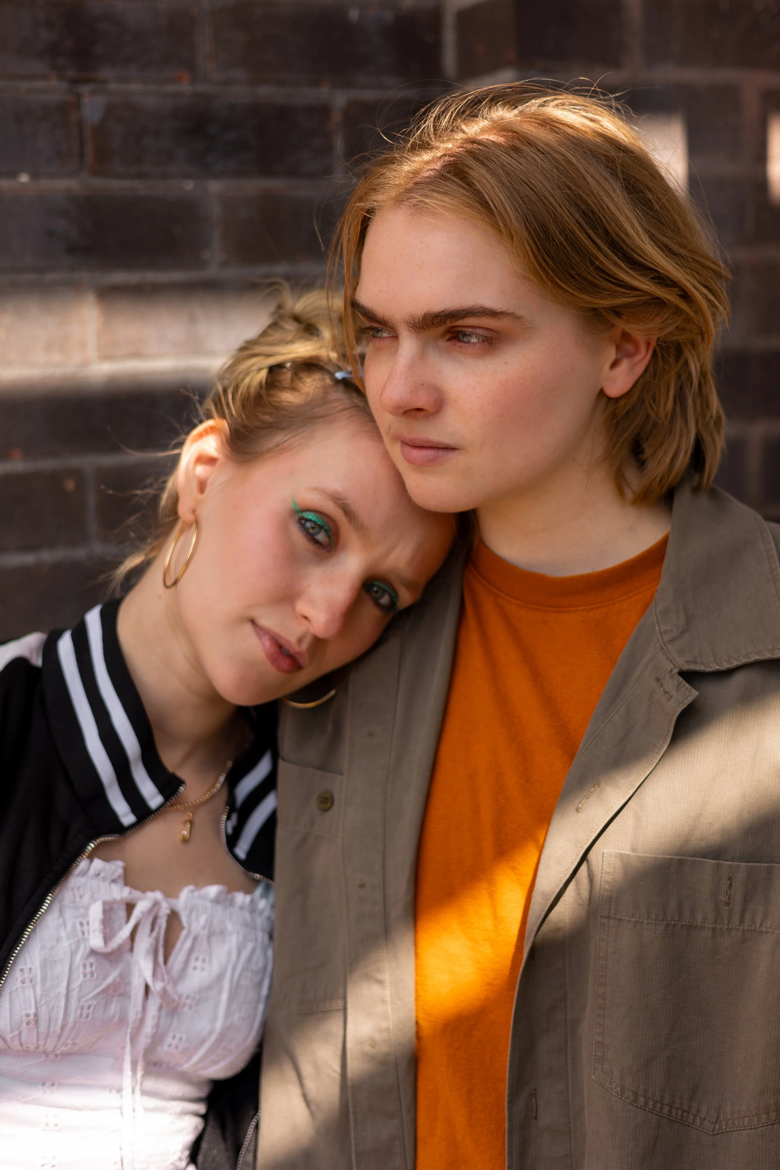 Romeo & Juliet photoshoot. A young woman, Madison Grace, with blue eyes and gold hoop earrings, leaning her head on the shoulder of a person, Izzie Harding-Perrott wearing an orange shirt and beige jacket. Photo by Lillie Sherry.
