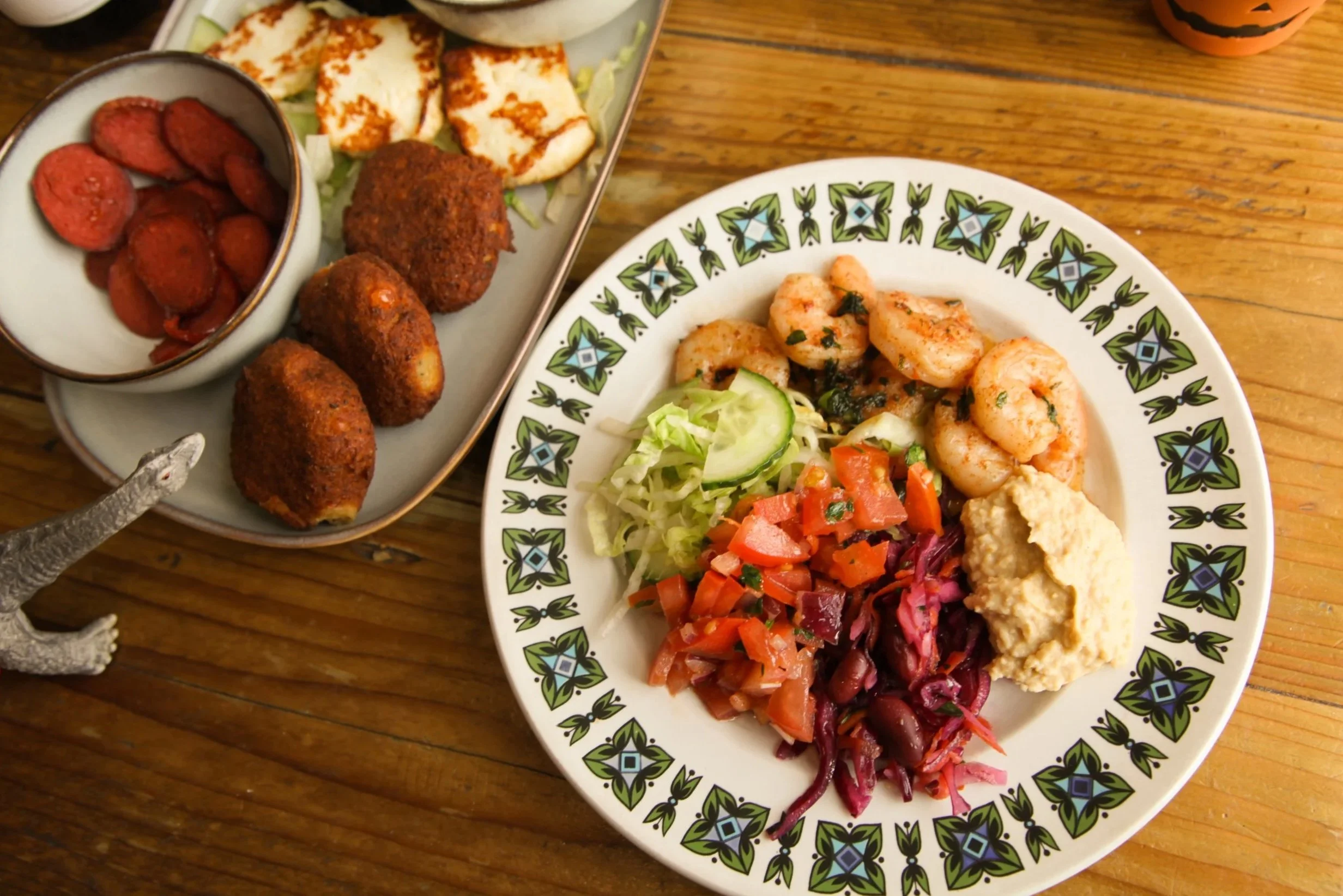 A spread of various foods on a wooden table, including a plate with shrimp and mixed vegetables, and a platter with falafel, grilled cheese, and roasted vegetables.