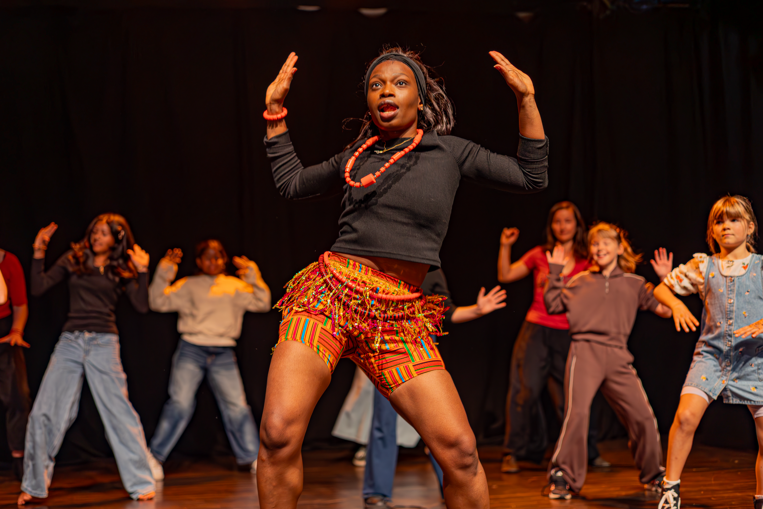 A woman dancing with her arms raised, wearing colorful traditional skirt, black long sleeve top, and red bead necklaces, on stage with children following her movements.
