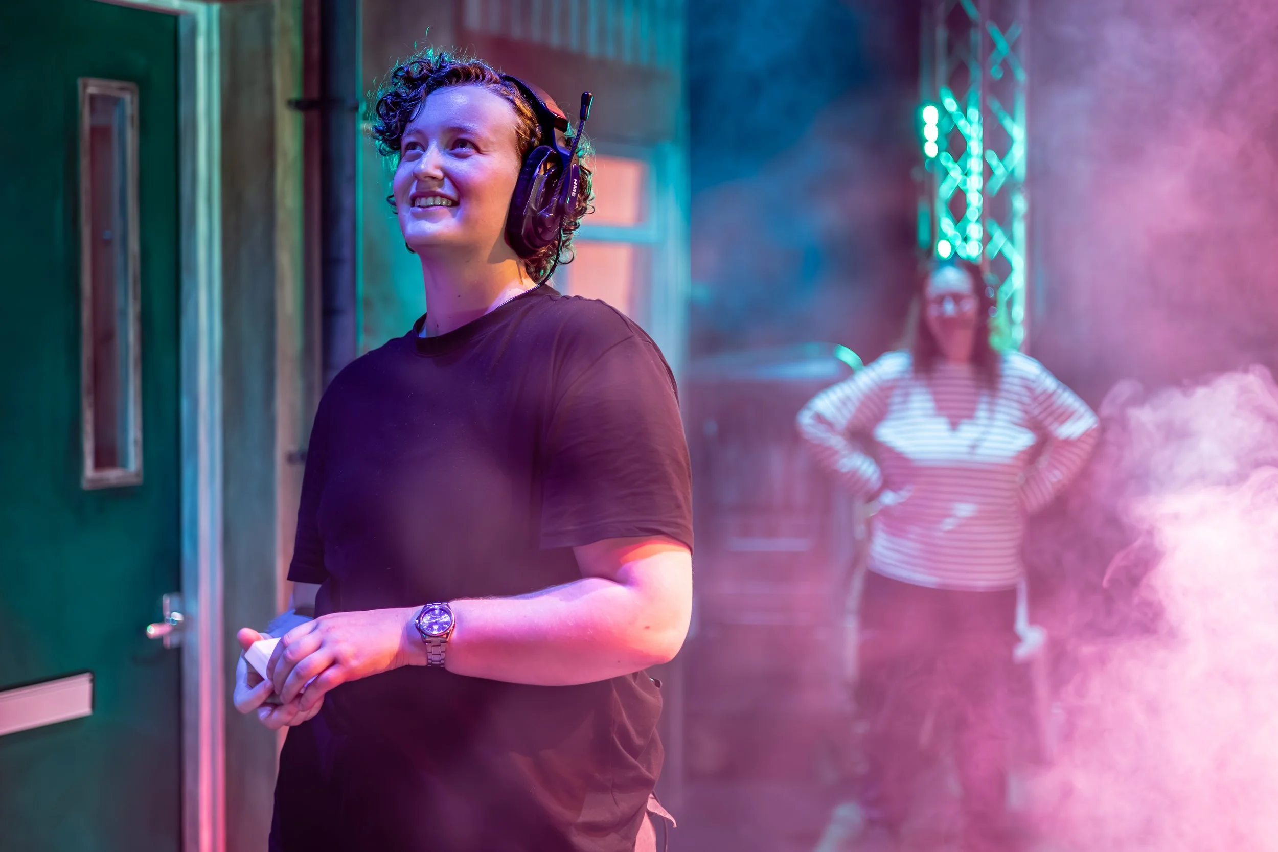 A woman with curly hair wearing headphones and a black shirt, smiling, standing in a room with colorful lighting and a person in the background.