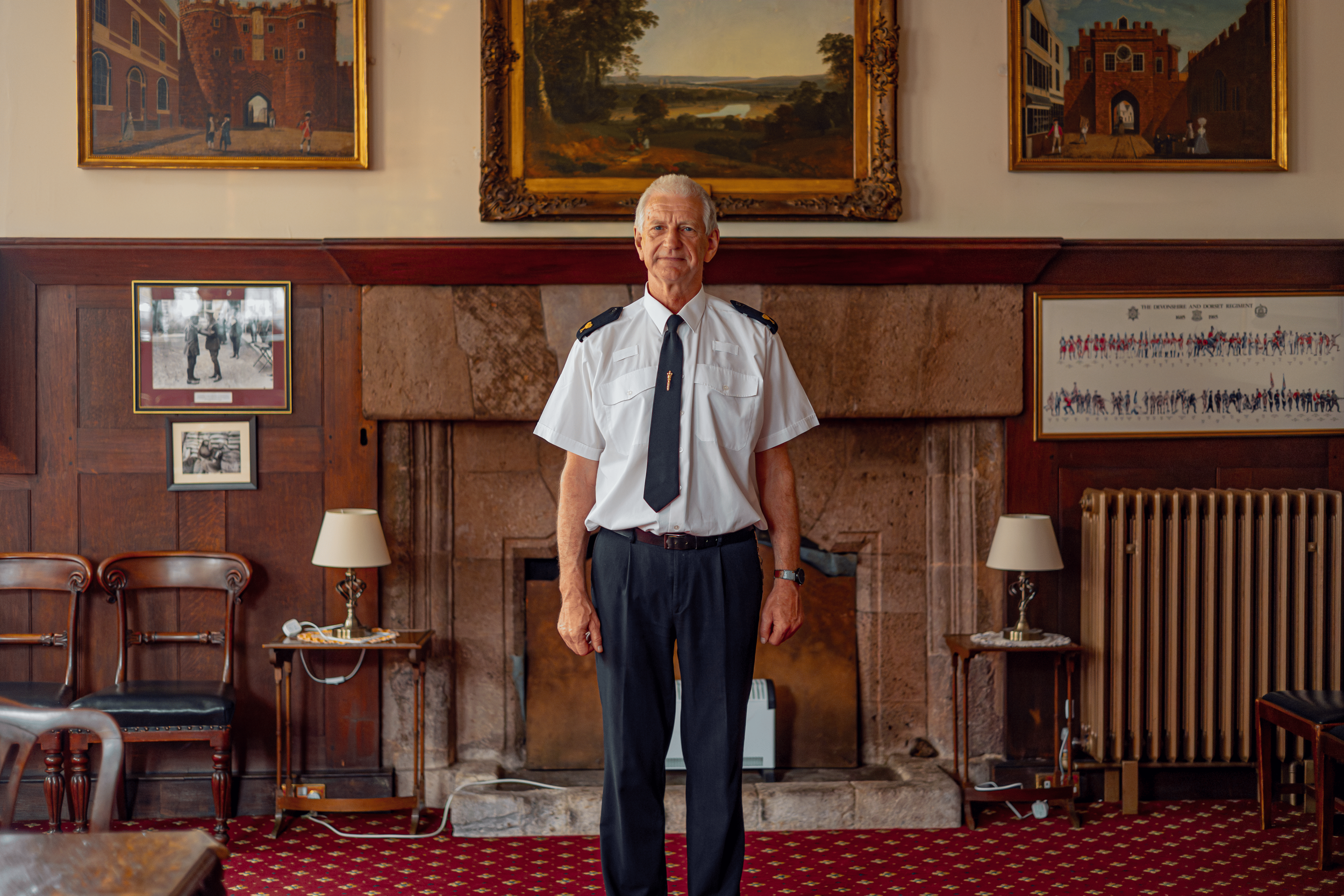 An elderly man in a white short-sleeved shirt and black tie stands in a wood-paneled room with framed paintings and photographs on the wall behind him. The room has a red carpet with a gold pattern, and a fireplace with a stone mantel. There are chai