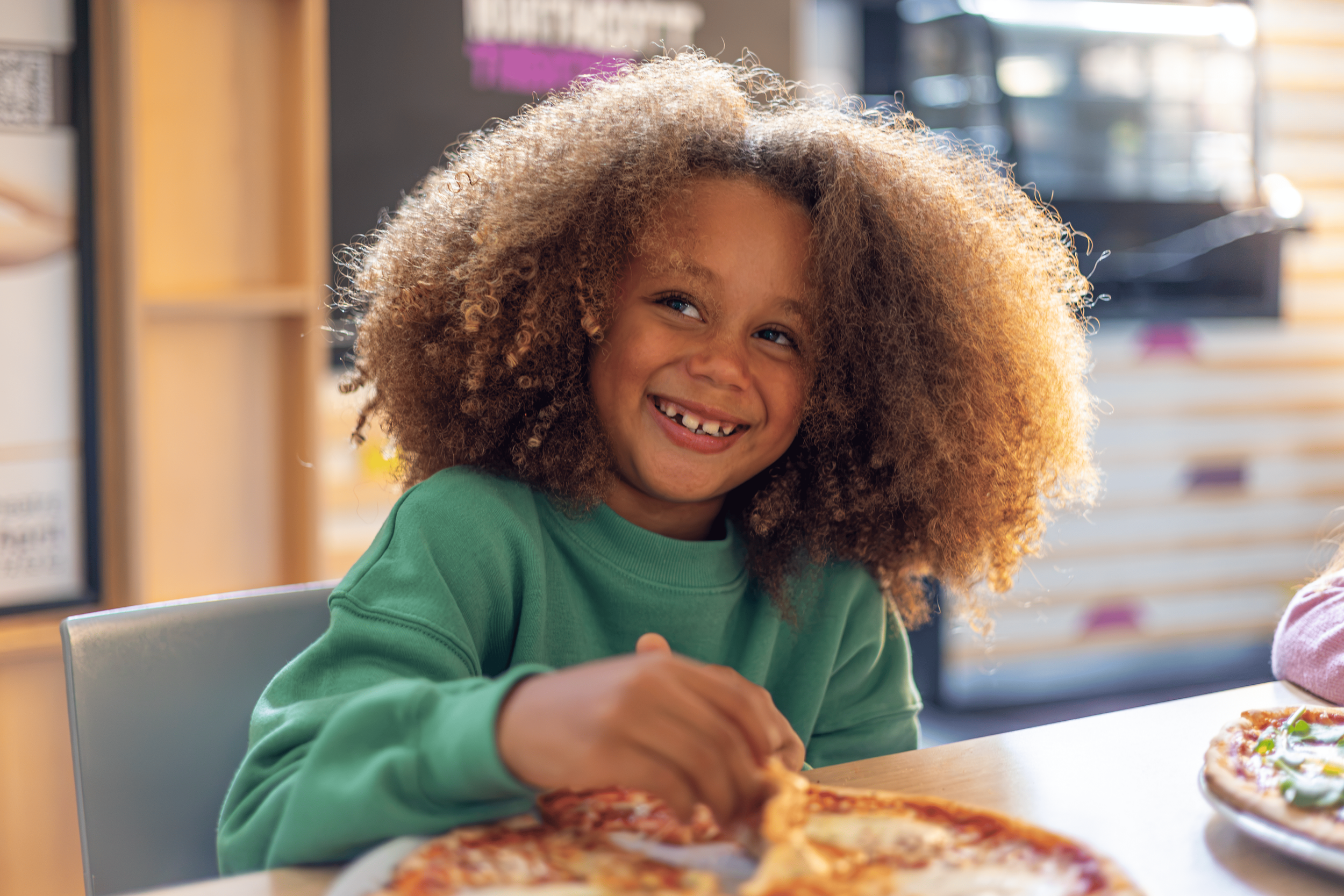 A young girl with curly hair, smiling and wearing a green shirt, sitting at a table with pizza in front of her in a restaurant.