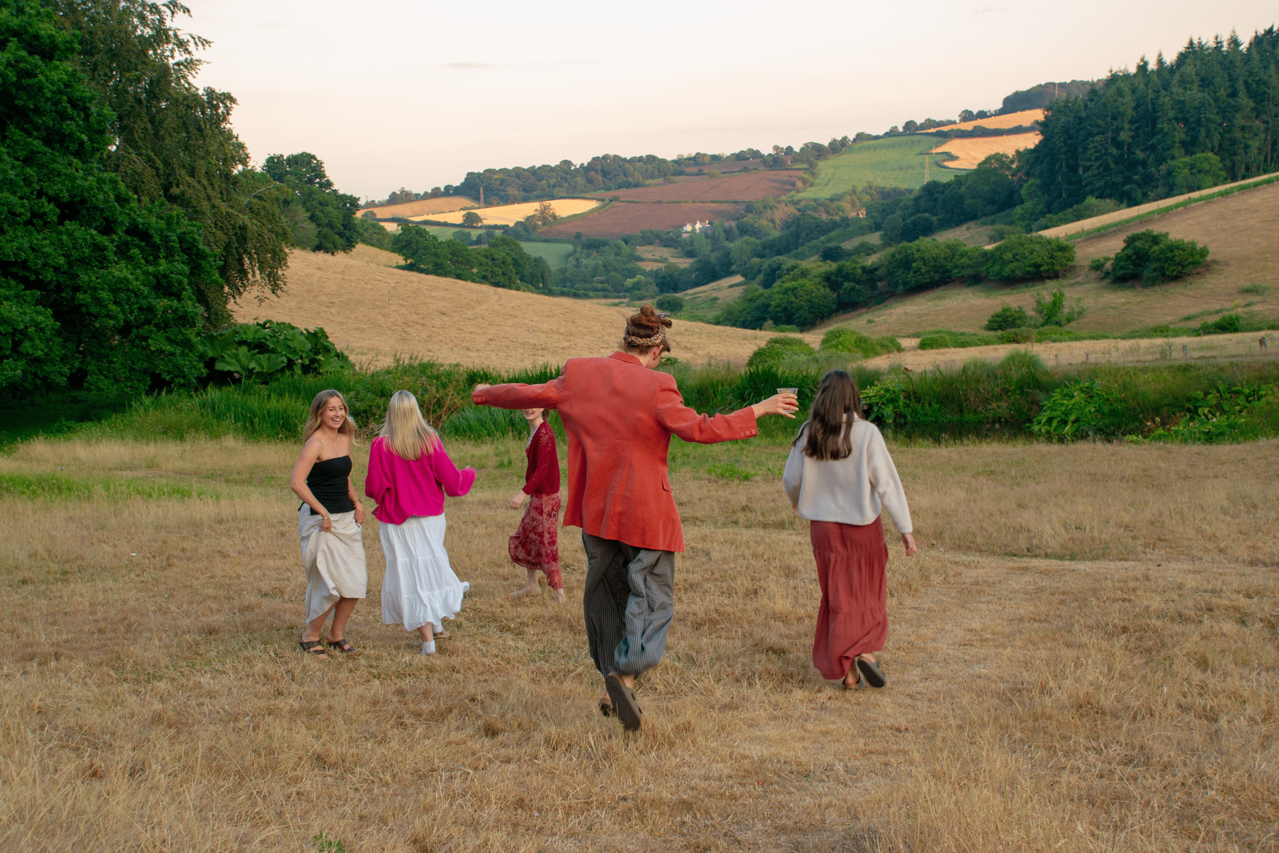 Group of seven young women and one man walking through a grassy field with rolling hills and patchwork farmland in the background. The man, who is in the center, is wearing a red jacket and has his arms outstretched, holding a glass.