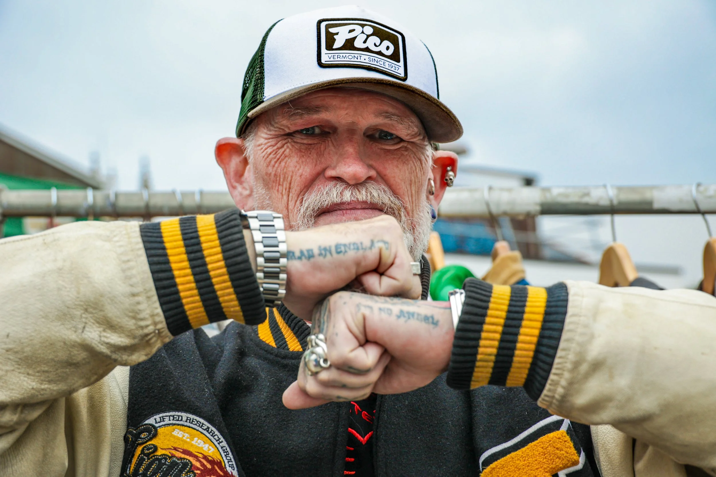 Close-up of a man with a beard and tattoos, wearing a Piklo trucker hat and a Harvard sweatshirt, holding a clothing hanger with his fists, standing outdoors under a cloudy sky. Photo by Lillie Sherry, Edinburgh-based photographer.