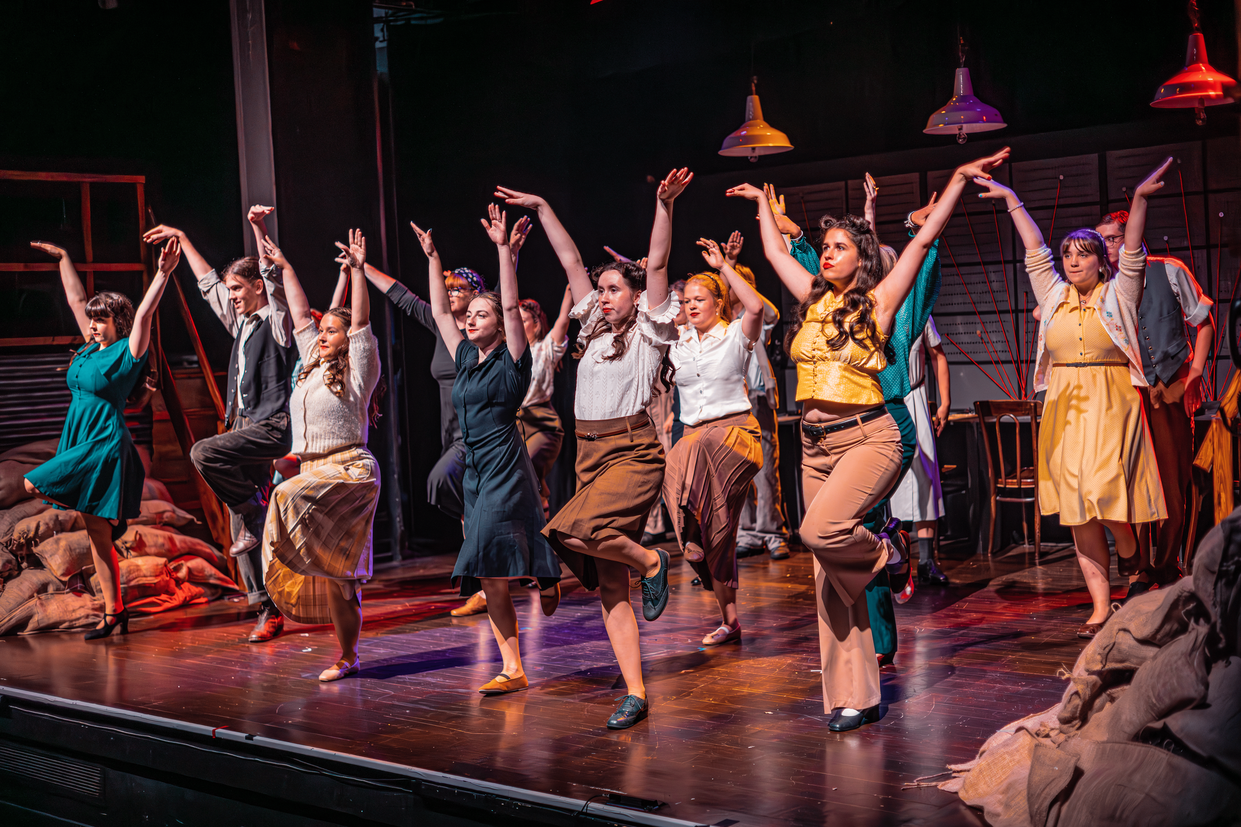 A group of actors are on stage performing a dance in vintage 1950s style clothing, with many raising their arms and smiling. Photo by Lillie Sherry, Edinburgh-based photographer.