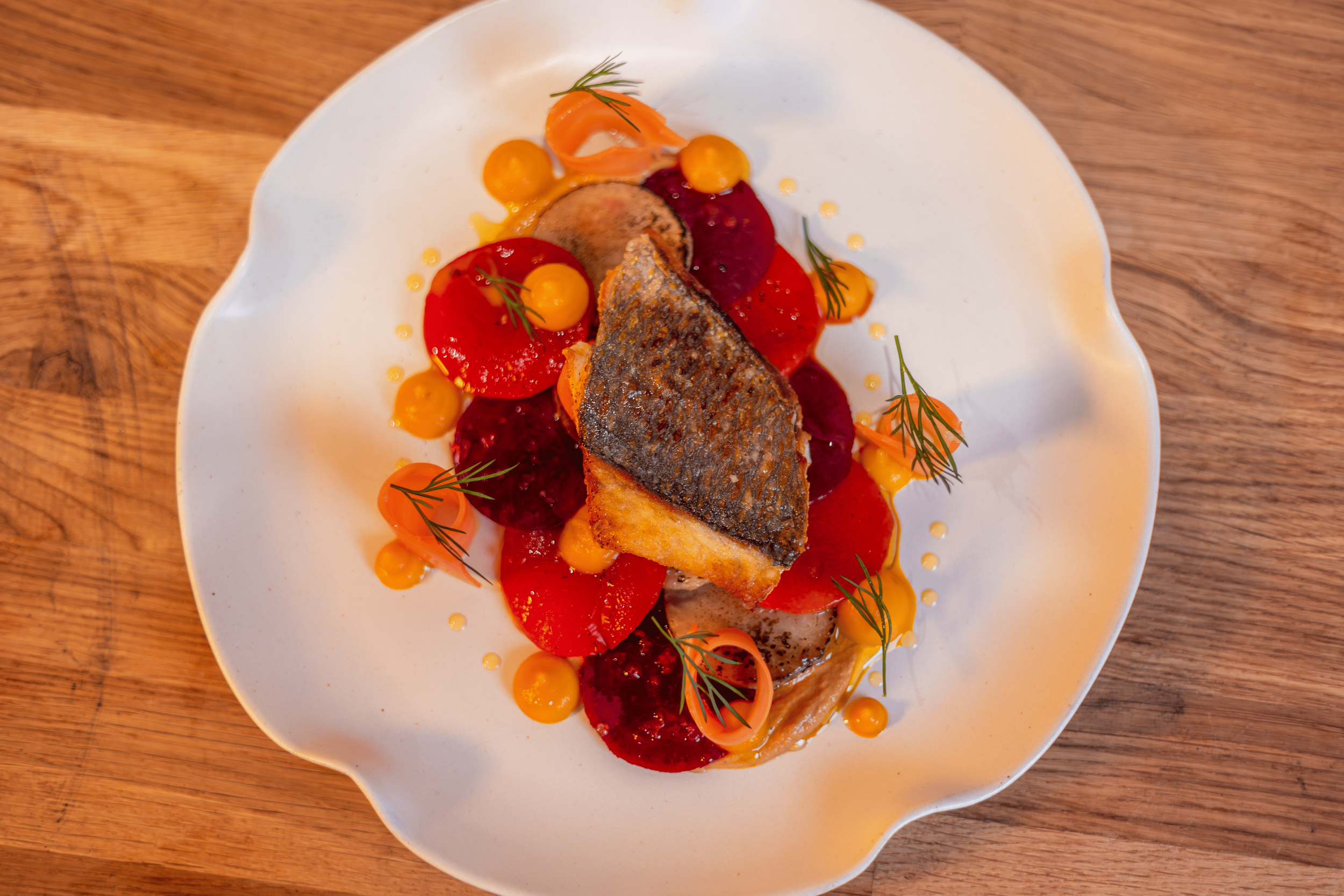 A plate of cooked fish with crispy skin, garnished with fresh herbs, and served with small yellow and red tomato-like vegetables, with a red sauce and yellow dots for decoration.