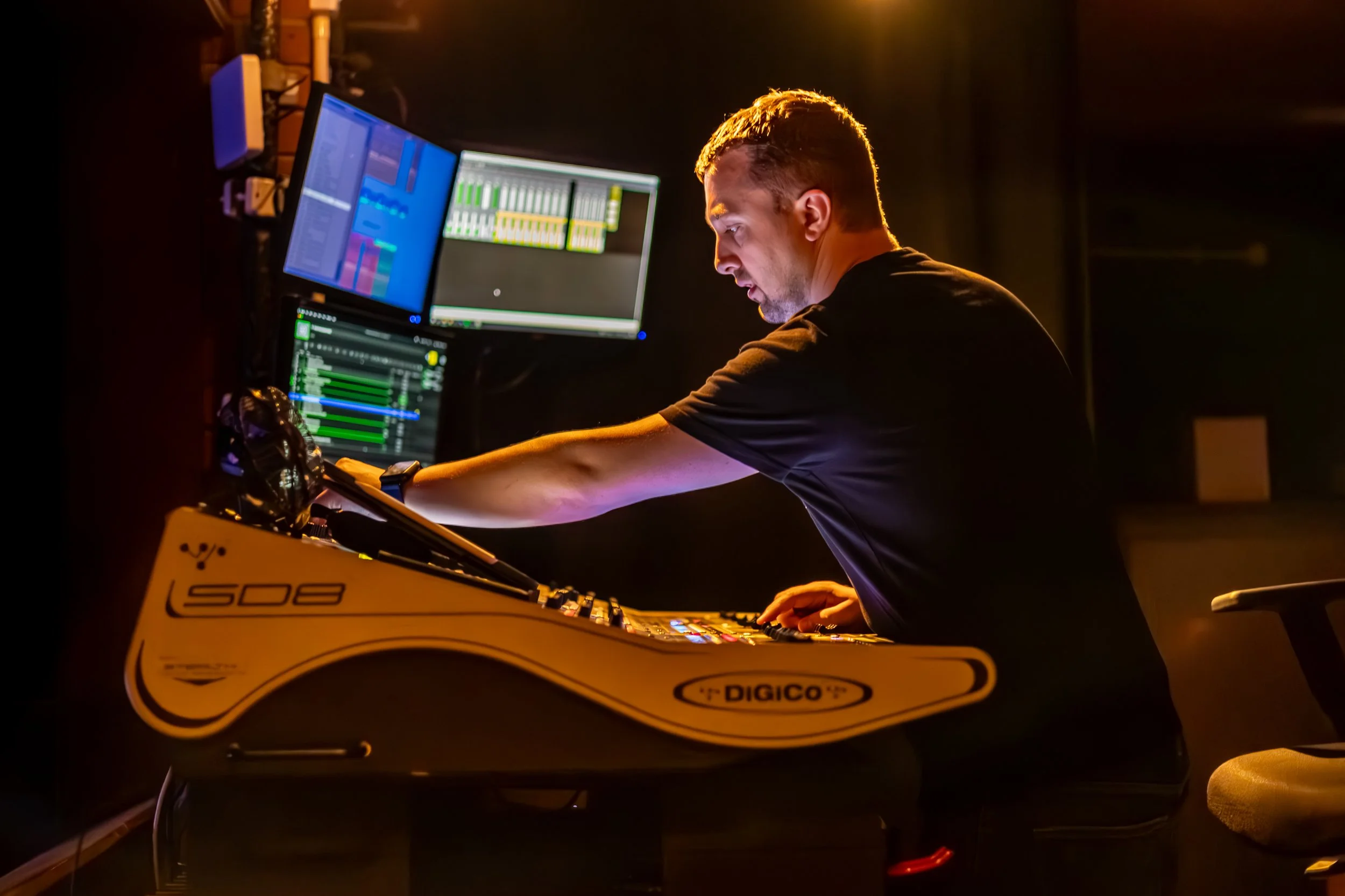 A man working at a music production studio with multiple monitors displaying audio editing software, sitting in front of a synthesizer, illuminated by warm lighting.