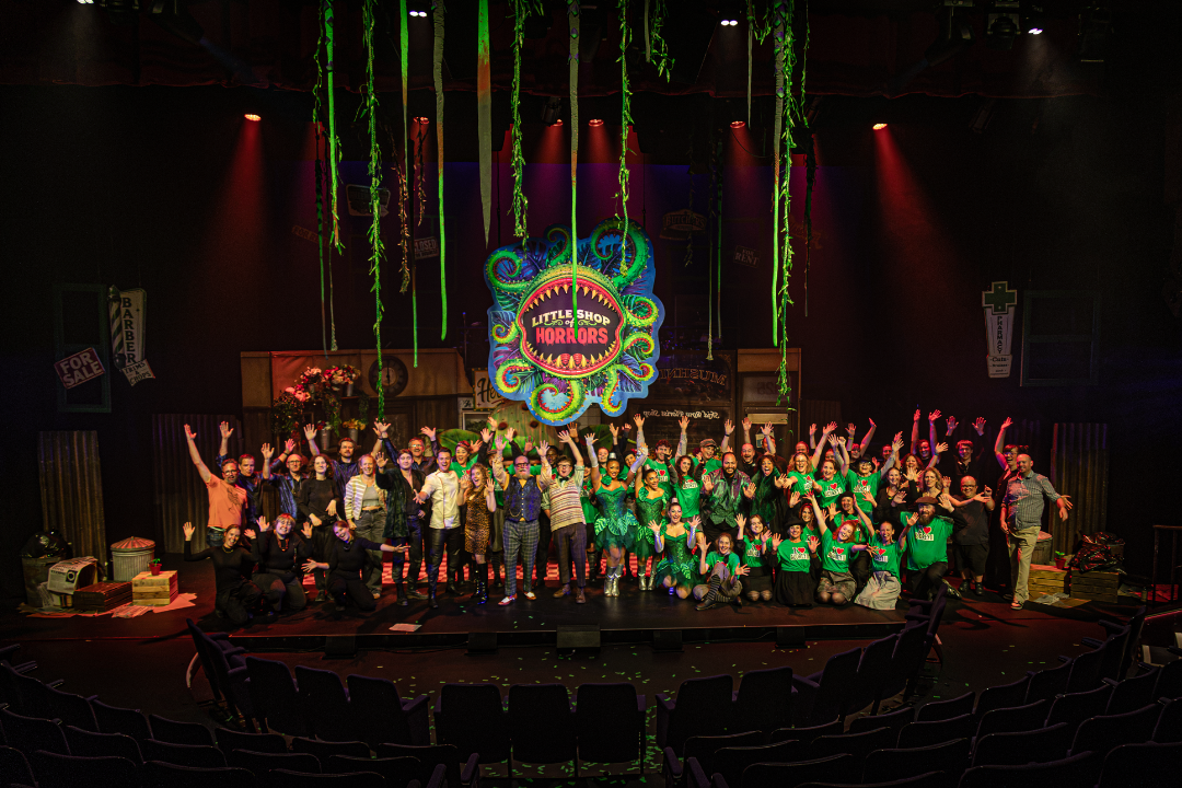 A large group of performers and crew members on stage at Little Shop of Horrors, with a colorful, whimsical backdrop and hanging green vines, celebrating their production.