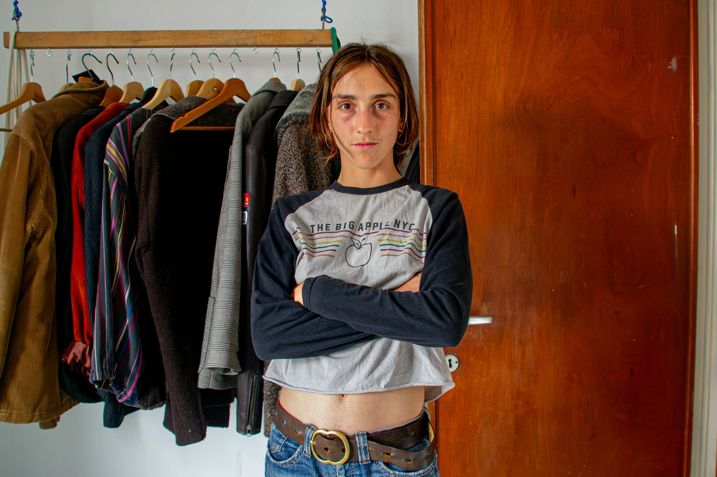 Young woman with short brown hair standing in front of a clothes rack with various jackets and shirts, crossing her arms, in a room with a wooden door to her right.