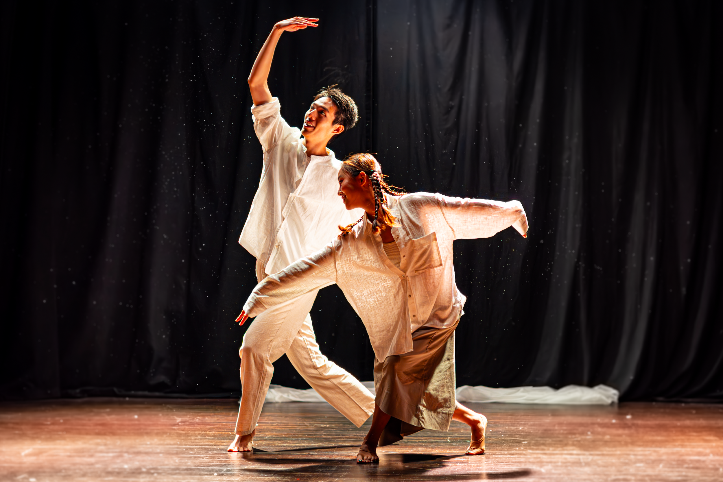 Two dancers performing on stage with black curtains and a dramatic spotlight. One dancer extends an arm upward, the other leans forward, both dressed in loose, light-colored clothing. Photo by Lillie Sherry, Edinburgh-based photographer.