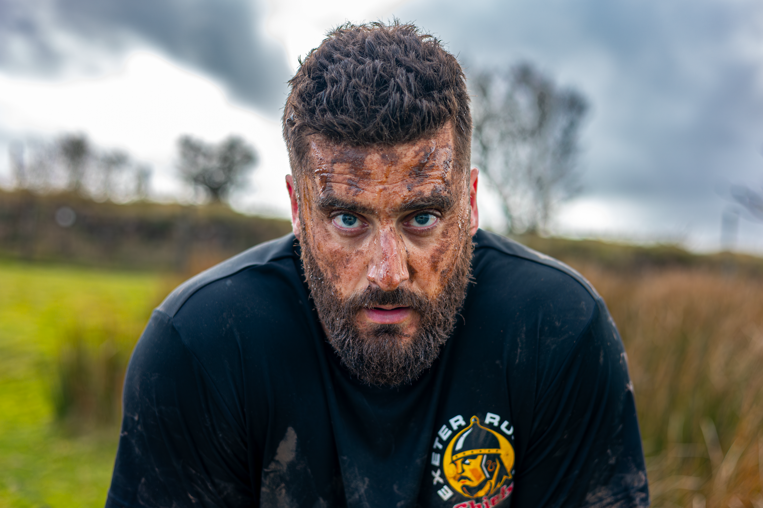 A man with wet, mud-covered face and bright blue eyes wearing a black shirt outdoors on a cloudy day.