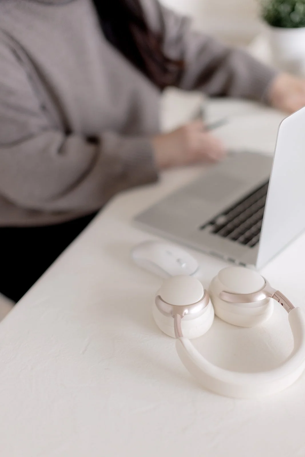 A pair of white over-ear headphones with pink accents resting on a white desk near a laptop and a computer mouse, with a person in the background working on a laptop.
