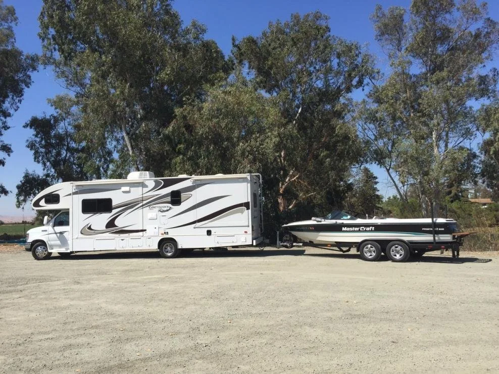 White RV camper with black and gray swooping graphics, attached to a black boat with the brand name 'MasterCraft,' parked on a gravel lot with trees and a blue sky in the background.