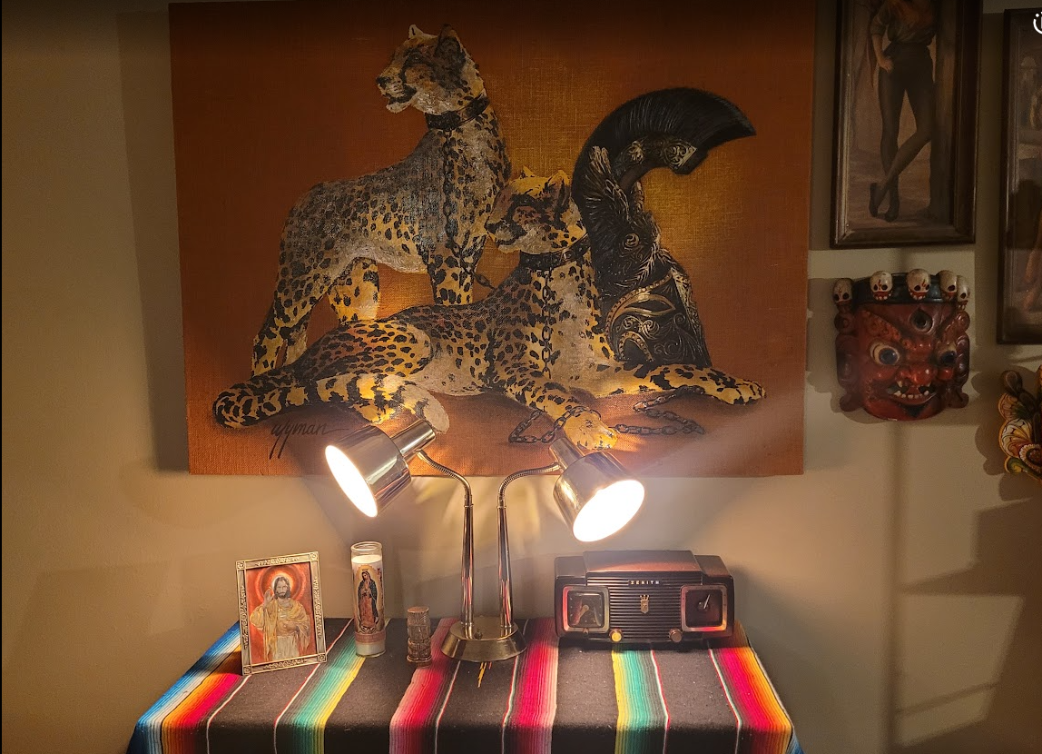 Eclectic altar display with leopard artwork, black feathered mask, red devil mask, religious icon, table lamp, vintage radio, and colorful striped blanket