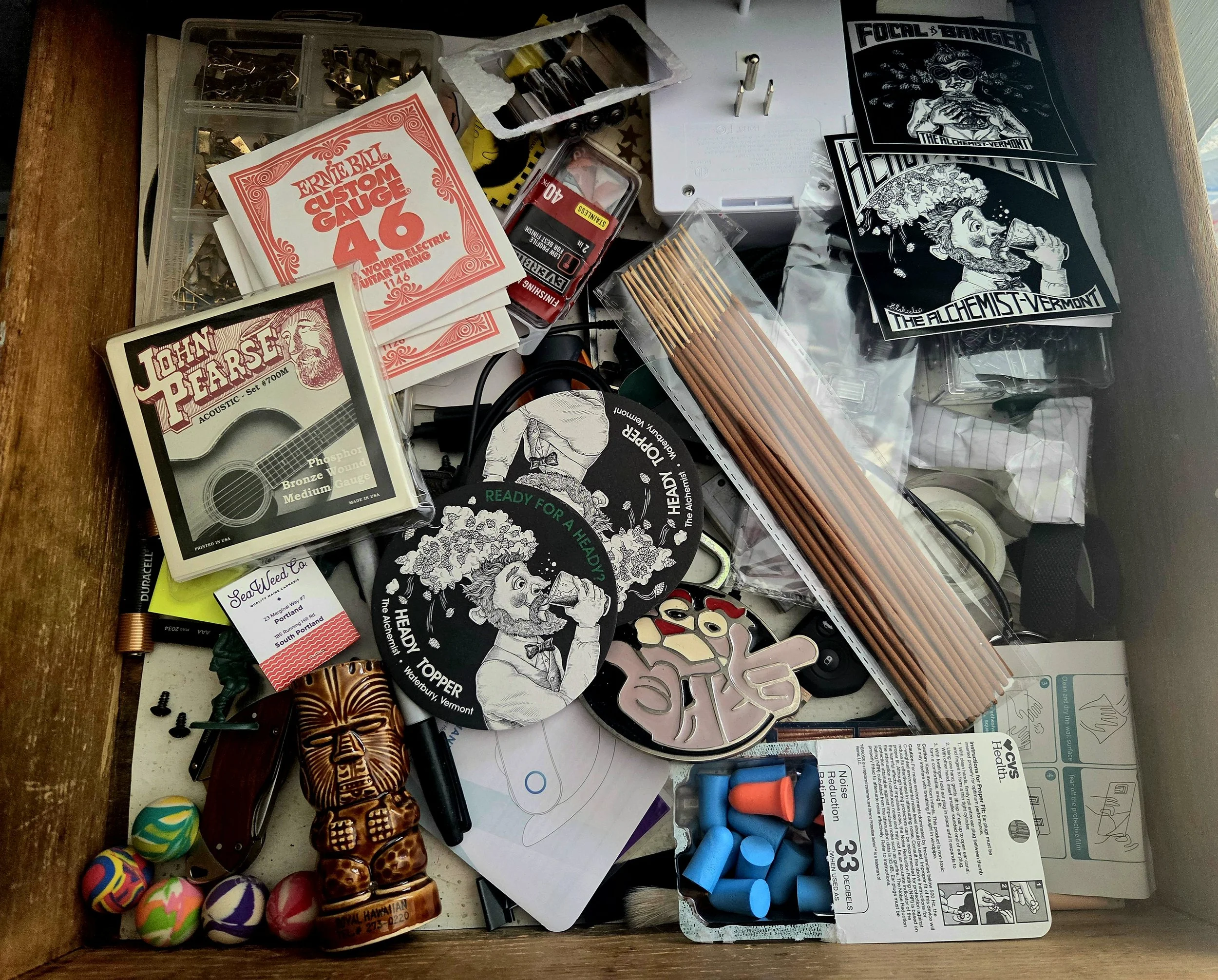 Overhead view of a cluttered workbench with guitar strings, sewing supplies, craft materials, stickers, earplugs, tools, and assorted small objects.