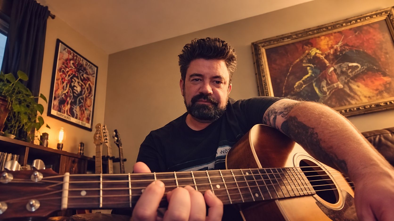 Bearded tattooed man playing an acoustic guitar in a warmly lit living room