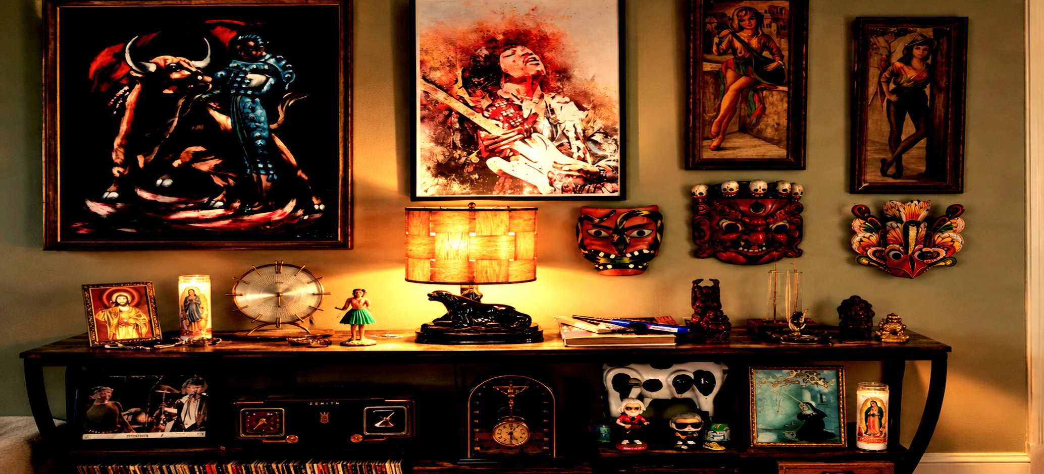 Eclectic vintage console display with framed artwork, colorful masks, religious candles, figurines, a table lamp, clock, and collectibles