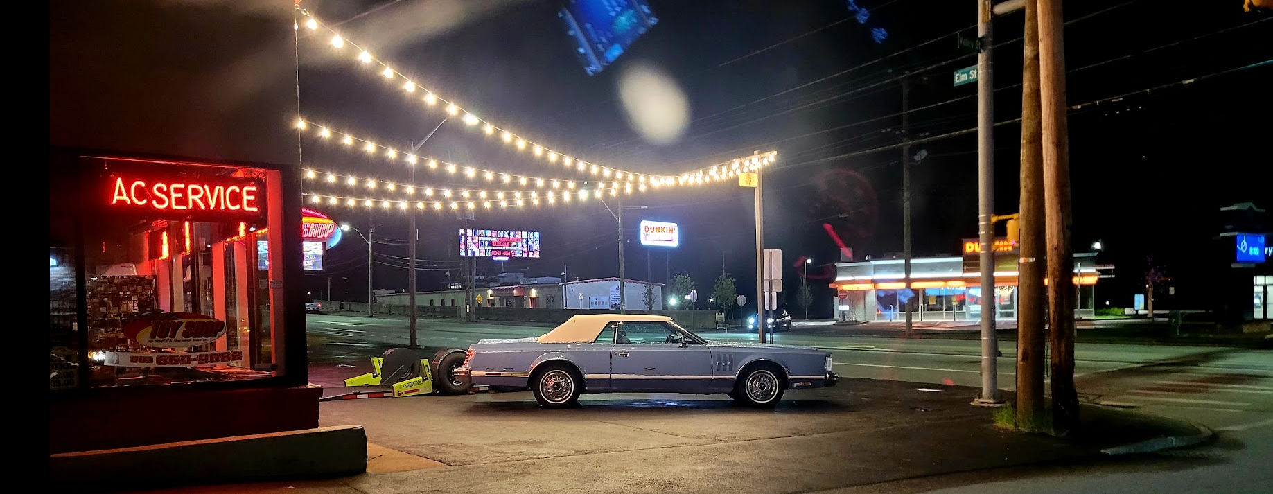 Classic blue convertible towing a small trailer outside a neon-lit auto service shop at night, with string lights and a Dunkin' across the street.