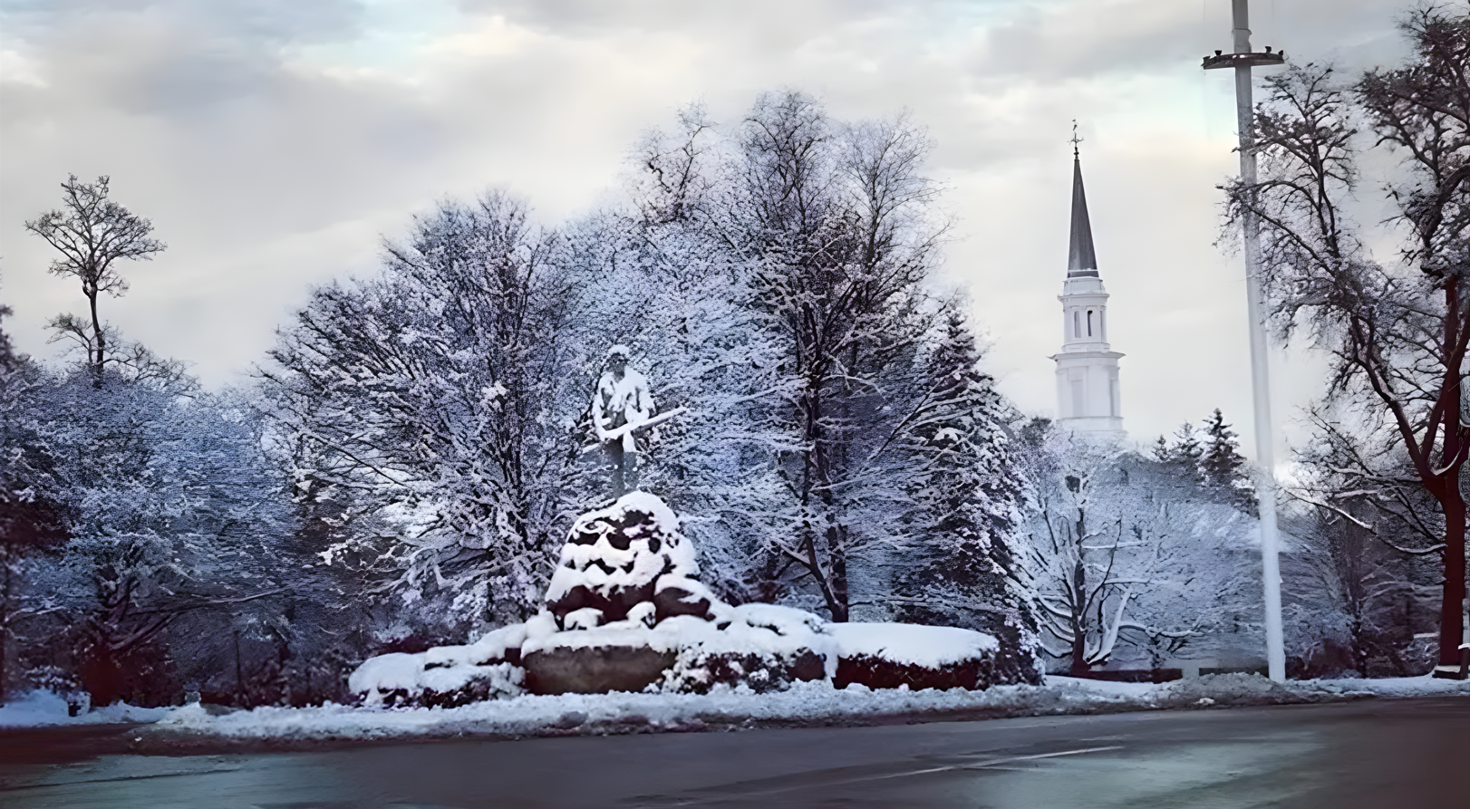 Snow-covered deer statue and trees in a park, with a white church steeple in the background.