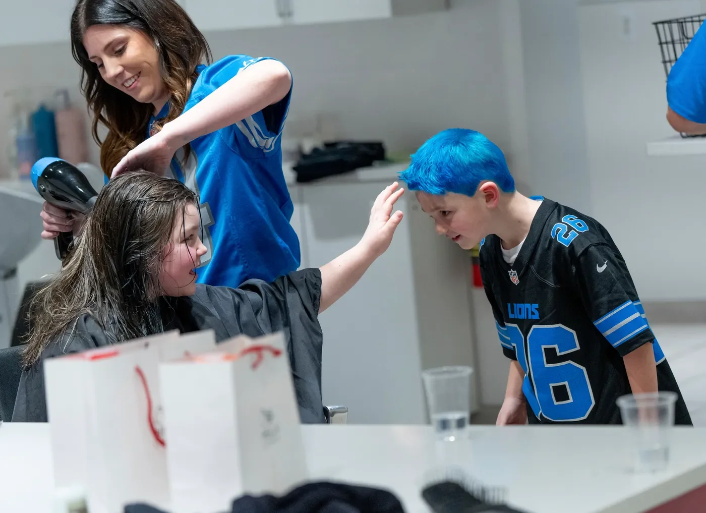 A woman with brown hair dries a young girl's hair with a hairdryer. The girl has wet hair and is sitting at a table in a salon or similar setting. A young boy with bright blue hair, wearing a black sports jersey with the number 26, is leaning forward and touching his head, looking at the girl and woman. There are white boxes and plastic cups on the table.