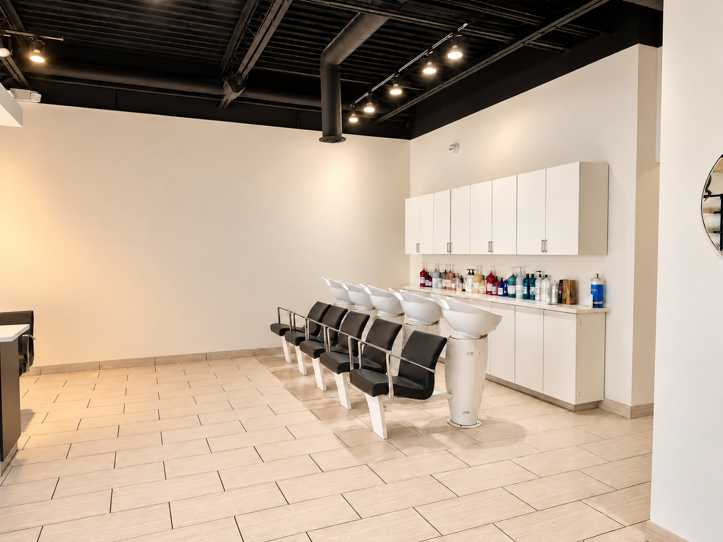 Hair salon with four black salon chairs facing white shampoo stations with various hair products on the counter and white cabinets on the wall. The space has a high black ceiling with track lighting and light wood tile flooring.
