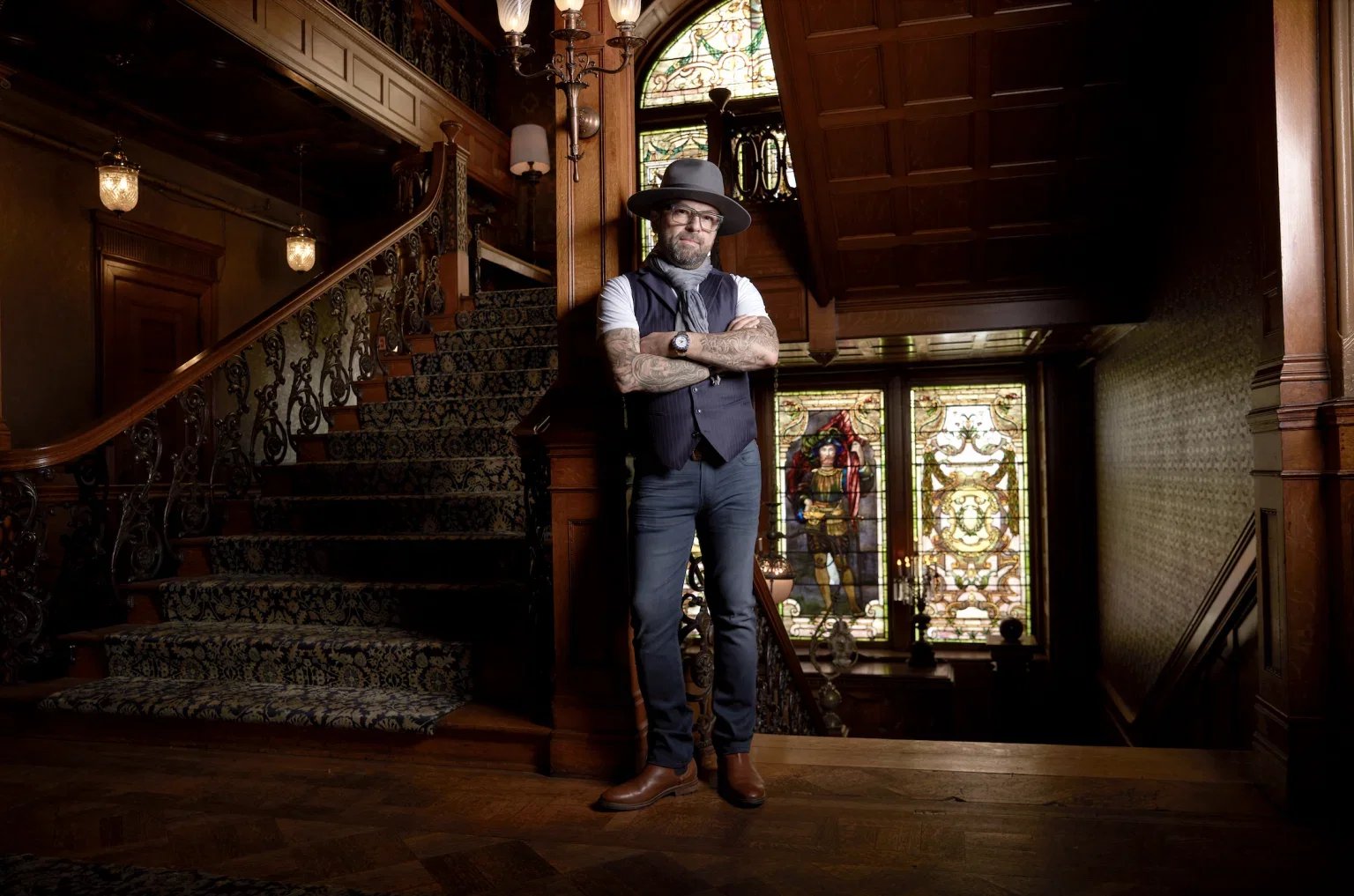 A man with tattoos, wearing glasses, a hat, a vest, and brown boots, standing with arms crossed in an ornate, vintage wooden staircase hall with stained glass windows.