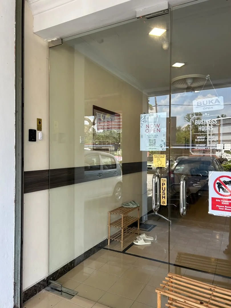 View of a glass storefront door with business hours, signage, and a 'Now Open' sign, showing shoes on the floor inside and parked cars outside.