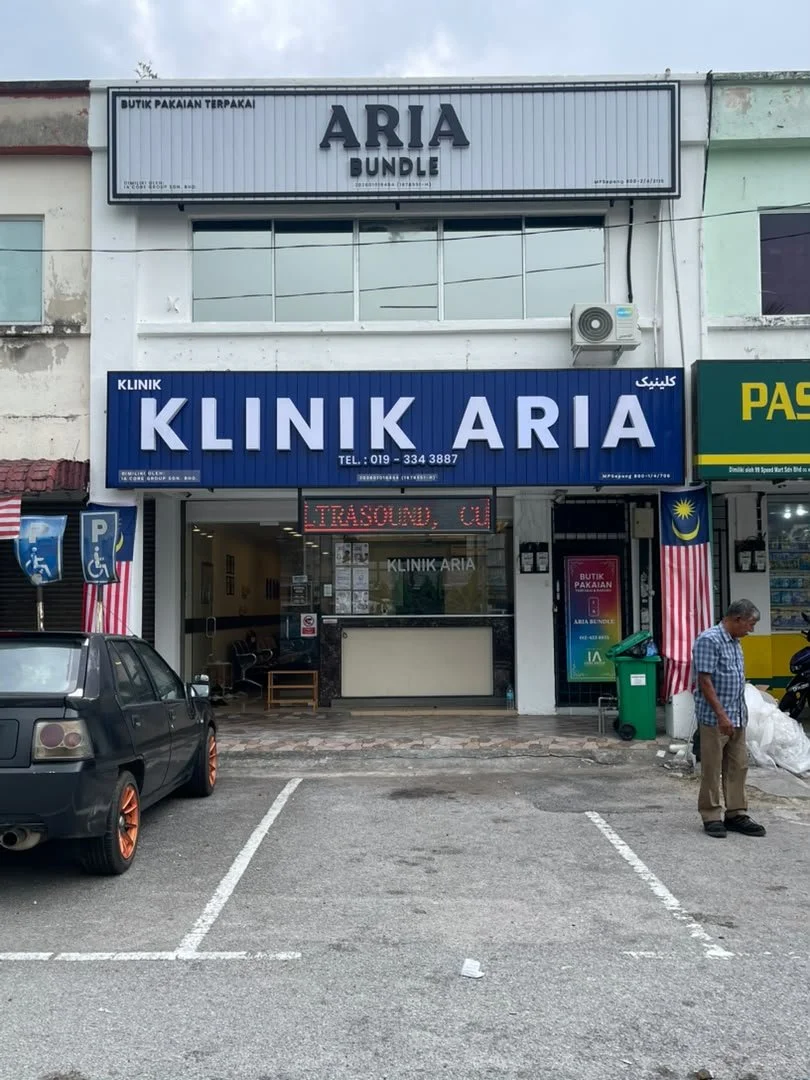 Front view of Klinik Aria, a clinic with a large blue sign, Malaysia flags on either side, parking spaces, a man standing outside, a black car, and various signs including a digital display in a city with partially visible storefronts.