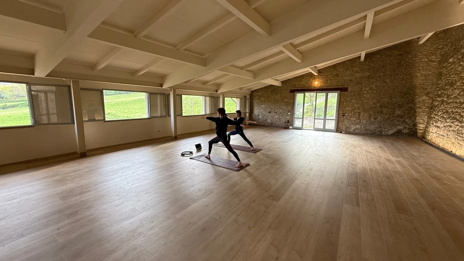 Two people practising yoga on mats inside the Limonaia at Castello di Galbino, with white beams and an exposed stone wall.