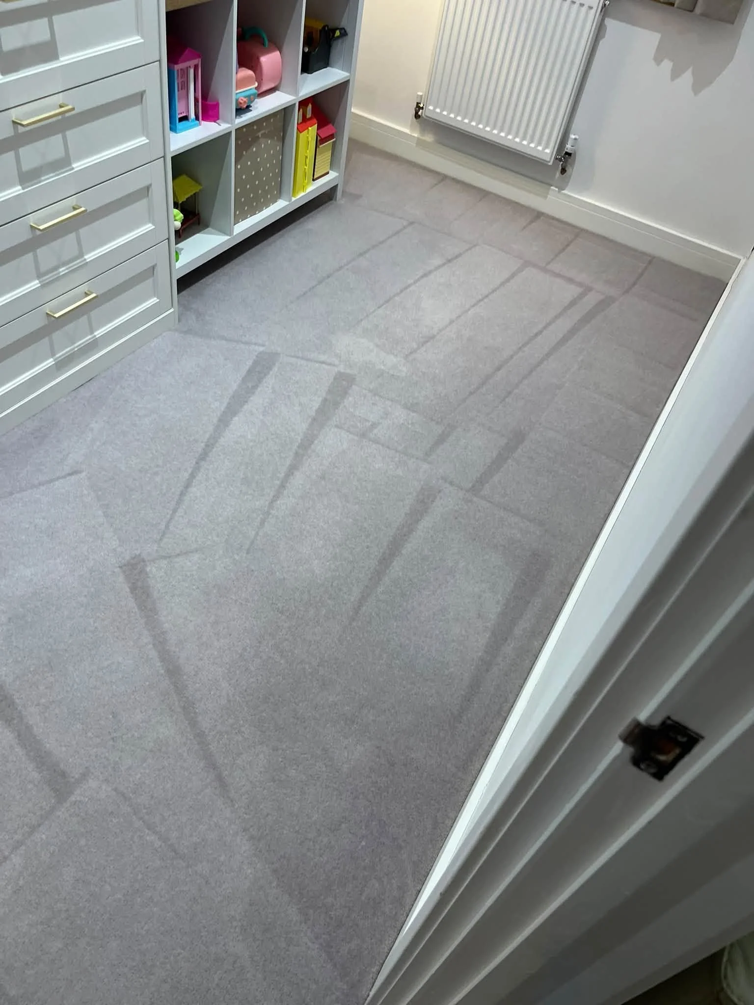 View of a room corner with light gray carpet, a white storage unit with drawers on the left, and a white radiator on the right wall.
