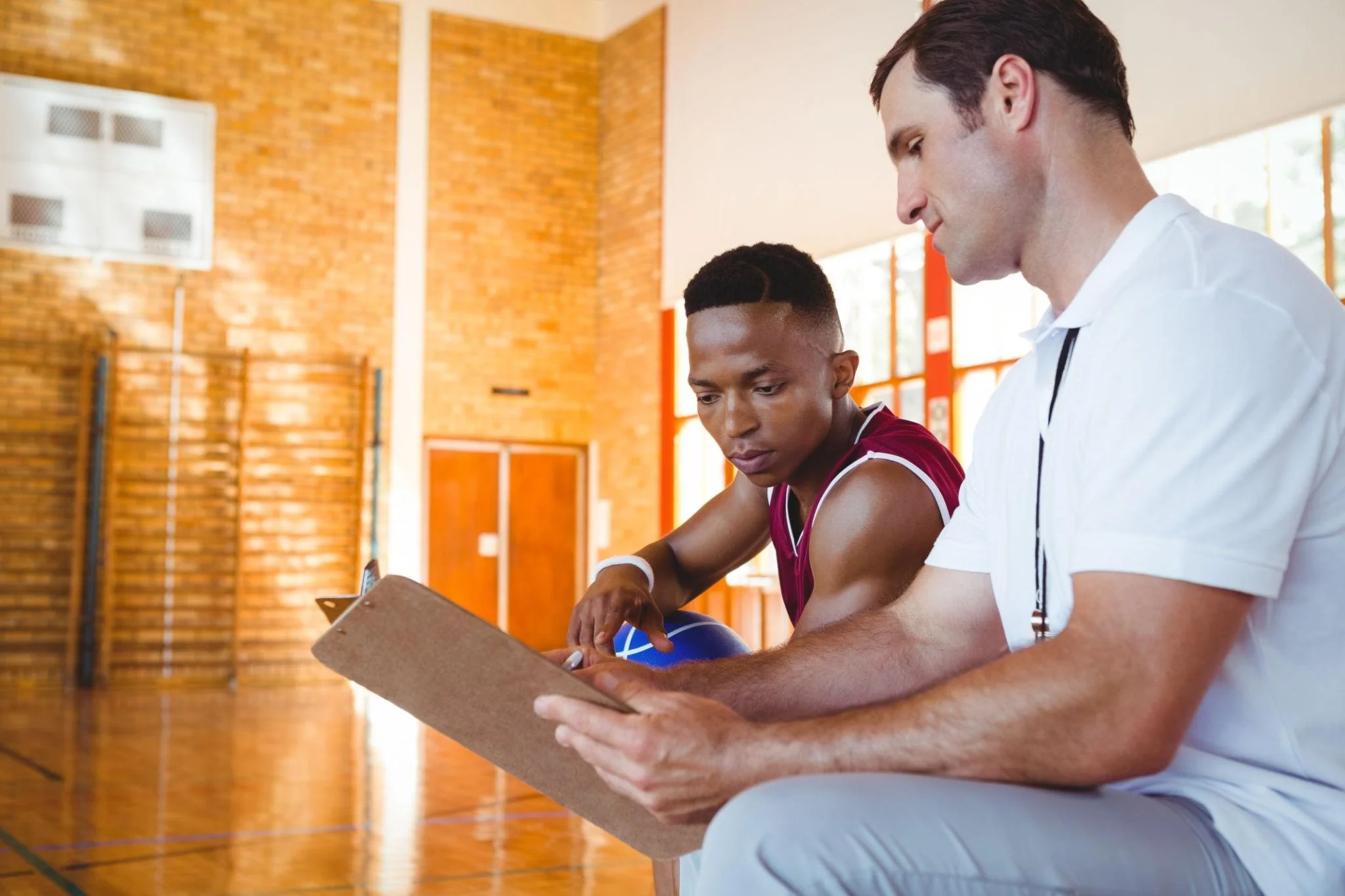 A male basketball player sitting on a bench with a coach in a gymnasium, looking at a clipboard.