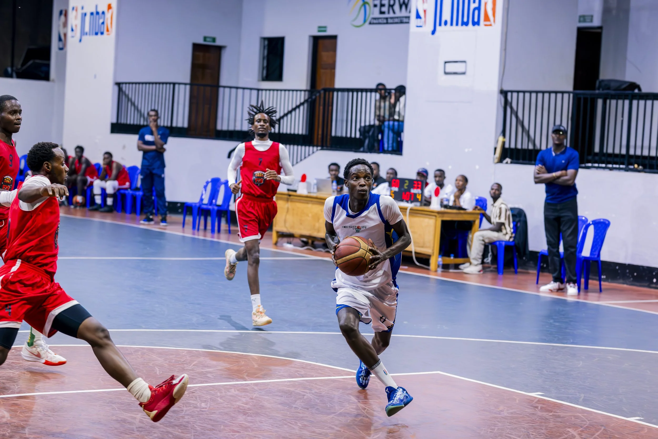 A basketball game with players running on the court; one player in a white uniform is holding the ball, while two players in red uniforms are nearby. People are sitting and standing watch from the sidelines, and a digital scoreboard is visible in the background.