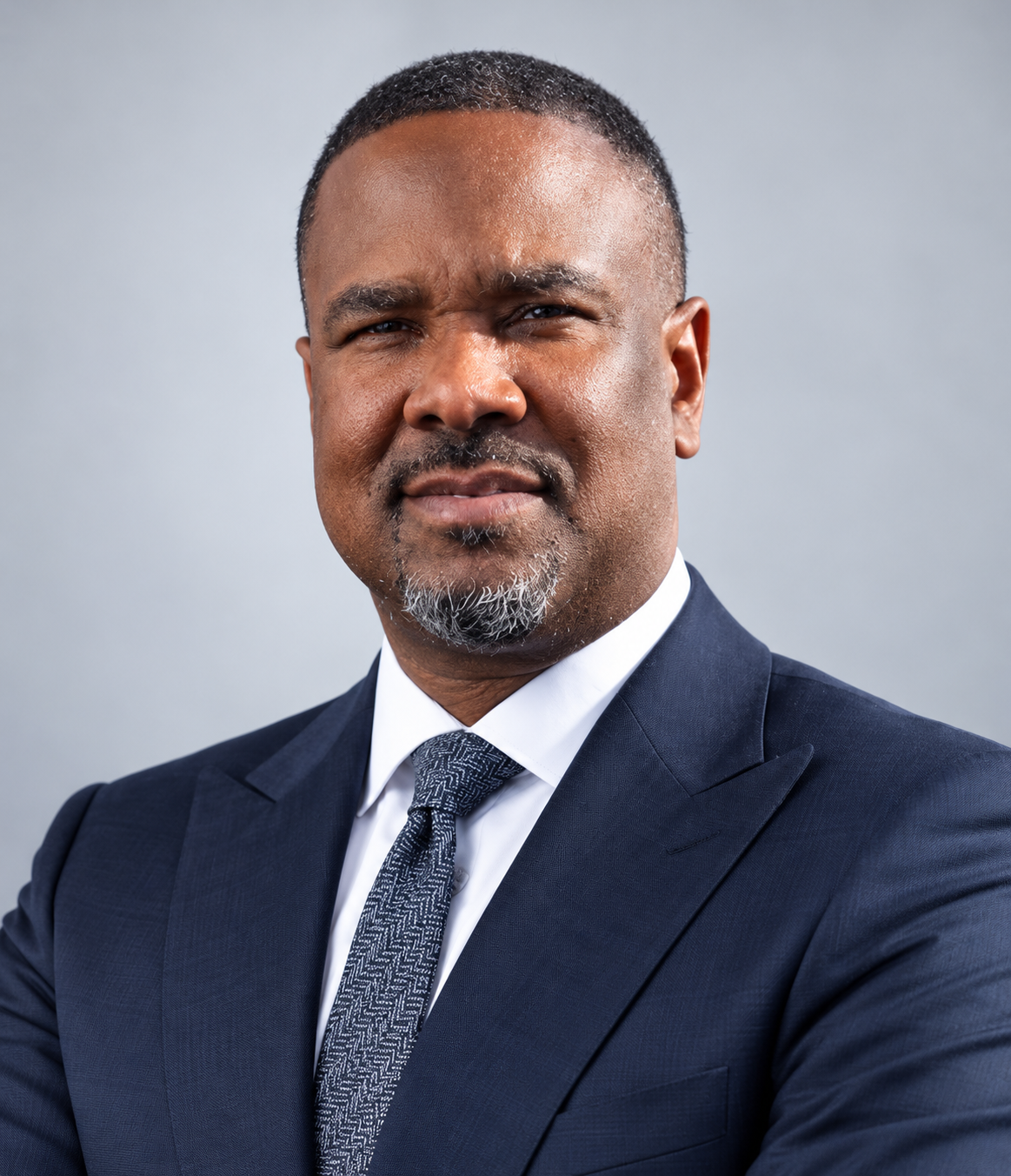 Professional portrait of a middle-aged man in a navy suit and patterned tie, with short hair, mild beard, and confident expression, against a plain gray background.