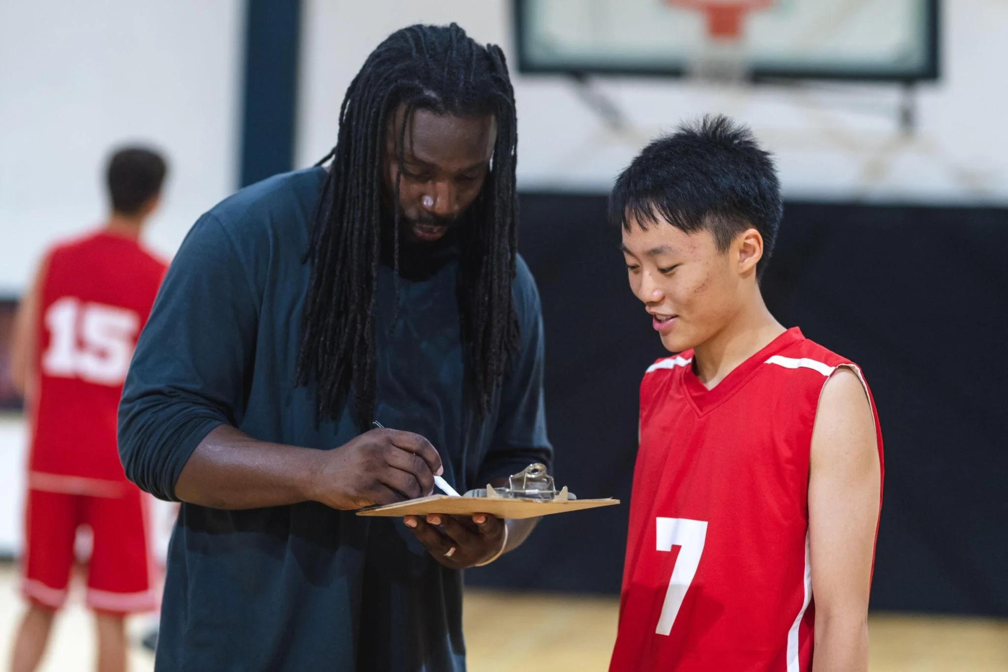 A coach signing an autograph for a young basketball player wearing a red jersey with the number 2 on it, in an indoor basketball court.