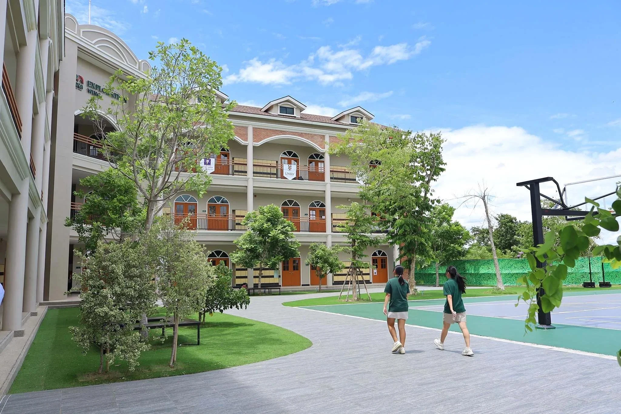 A school courtyard with green trees, a basketball court, and students walking. A large building with arched windows and balconies is in the background, with a partly cloudy sky overhead.