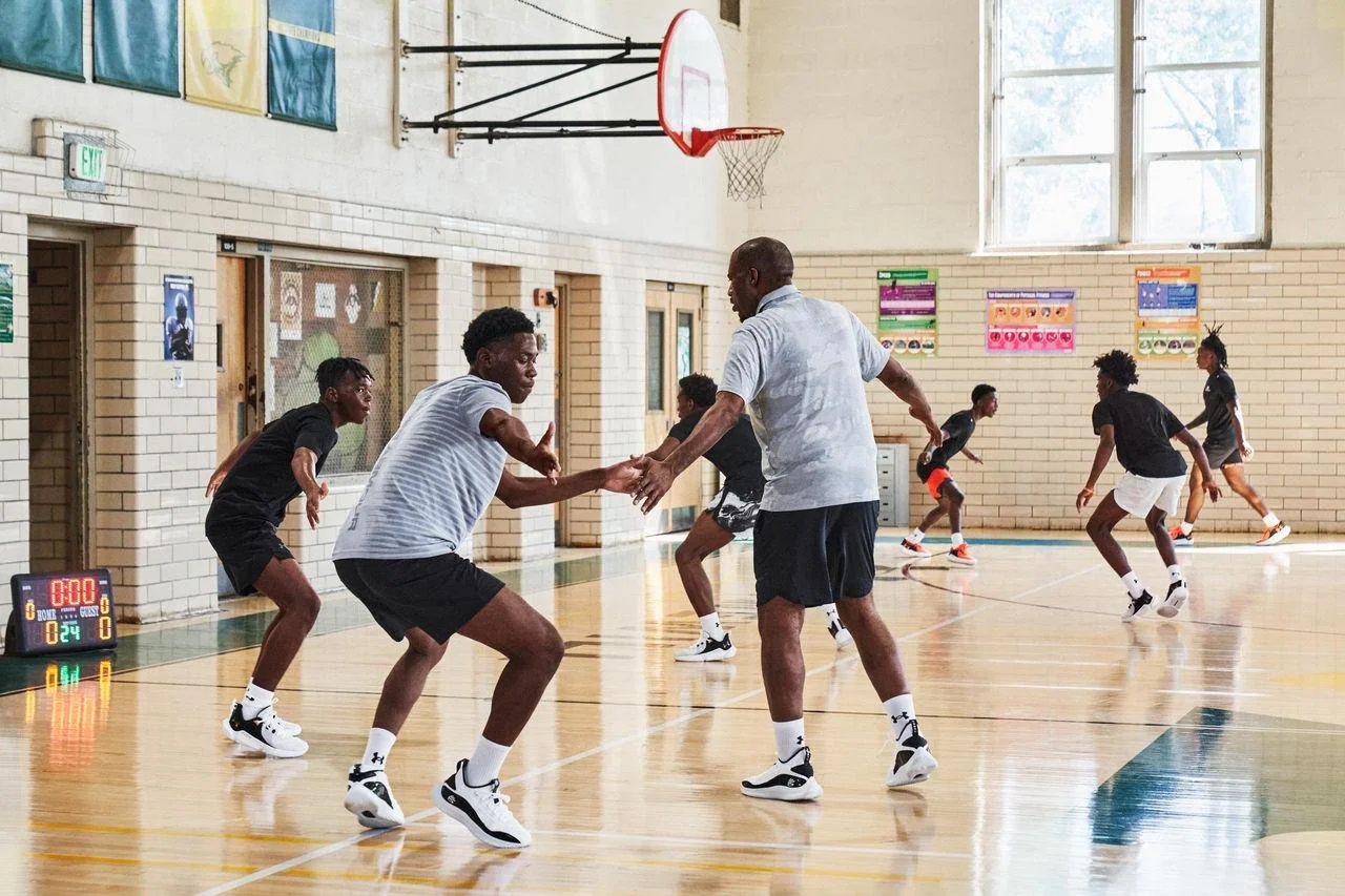 A group of young basketball players practicing in a gym under the supervision of a coach, with some players actively stretching and running.