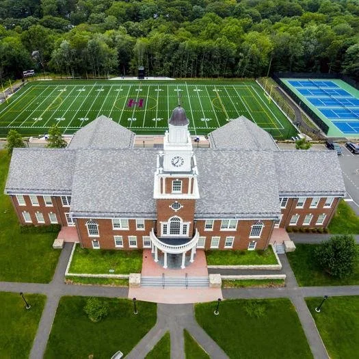 Aerial view of a large brick college building with a clock tower, front steps, and surrounding green lawn. Behind the building, there are football and tennis fields.